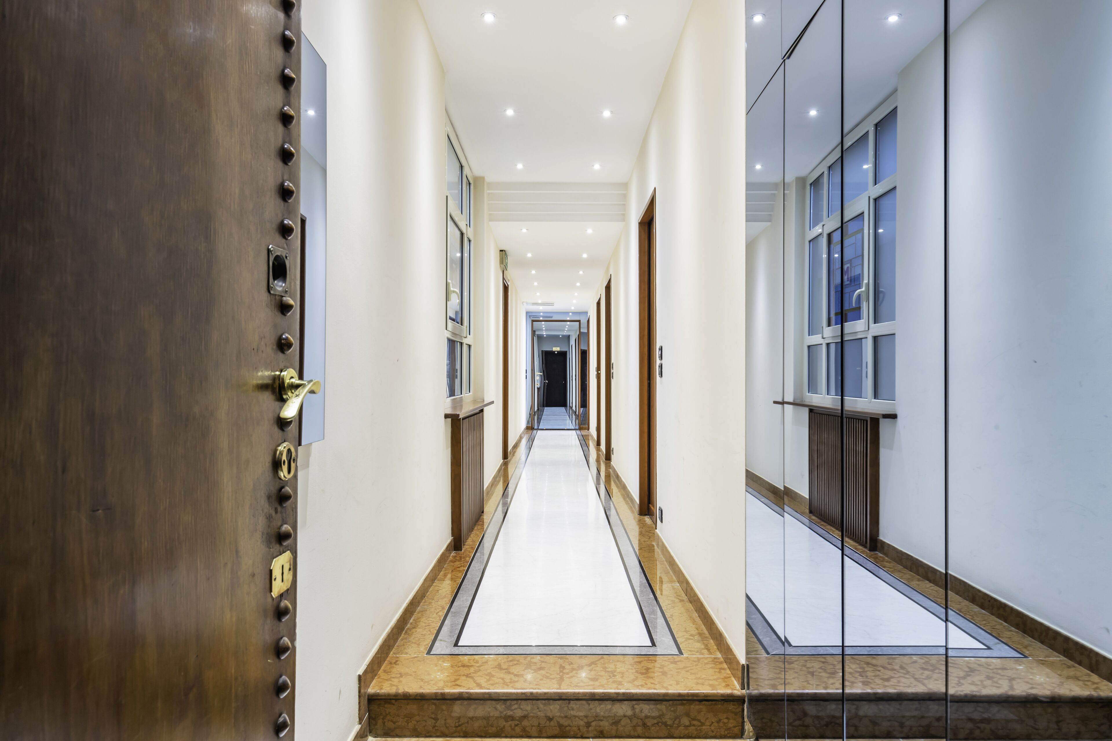A long, narrow hallway with a marble floor, wooden doors, and a glass wall on the right.