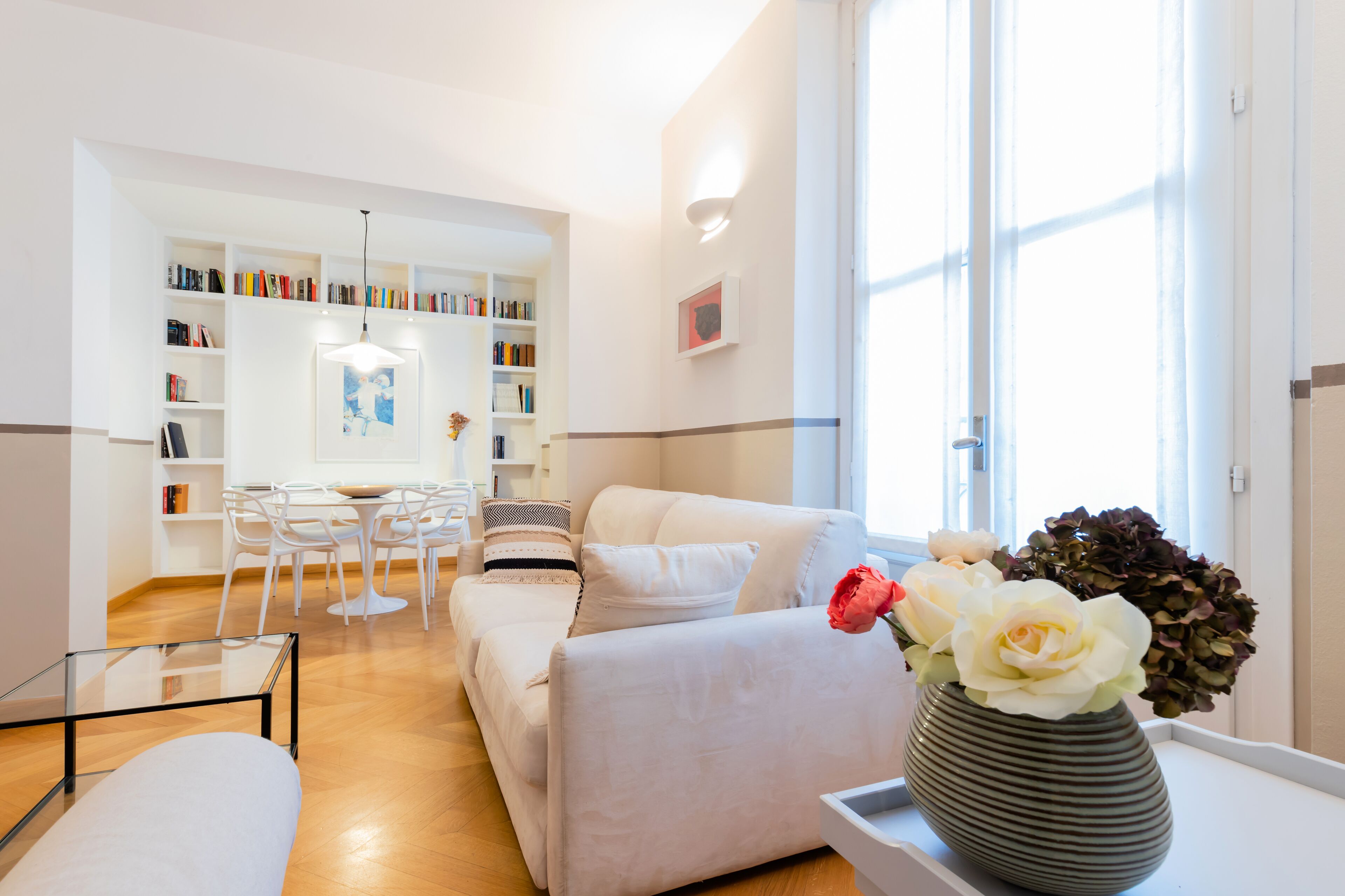 A modern living room with a sofa, a glass coffee table, and a dining area with a white table and chairs. There is a bookshelf filled with books and a vase with flowers on a side table.
