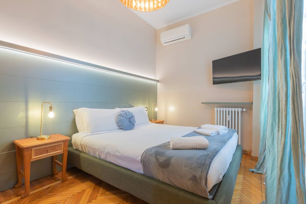 A modern hotel room with a large bed, a bedside table with a lamp, a flat-screen TV mounted on the wall, and a radiator.