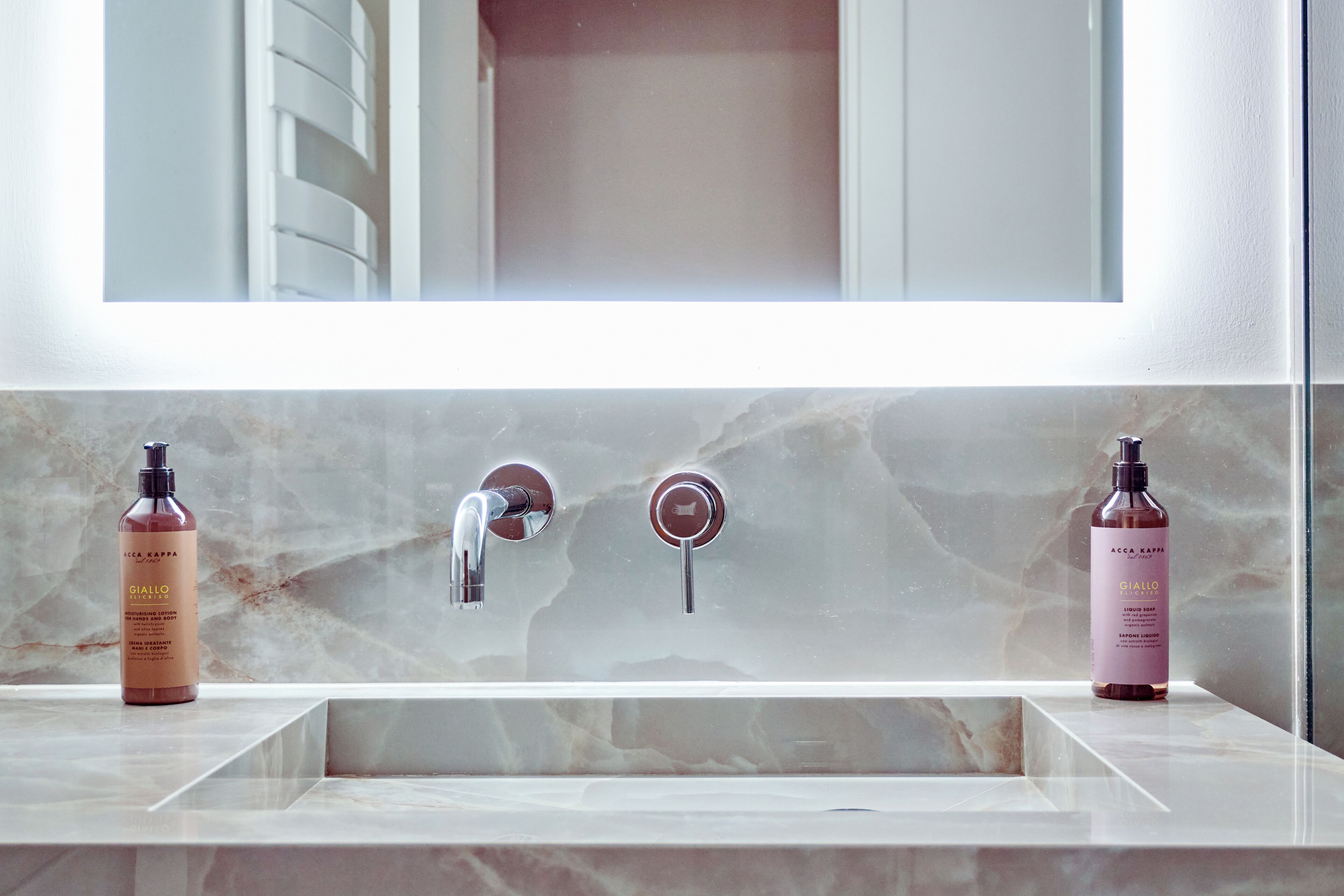 A bathroom with a marble sink, two bottles of A.C.C.A Kappa Giallo Elisirso liquid soap, and a chrome faucet.