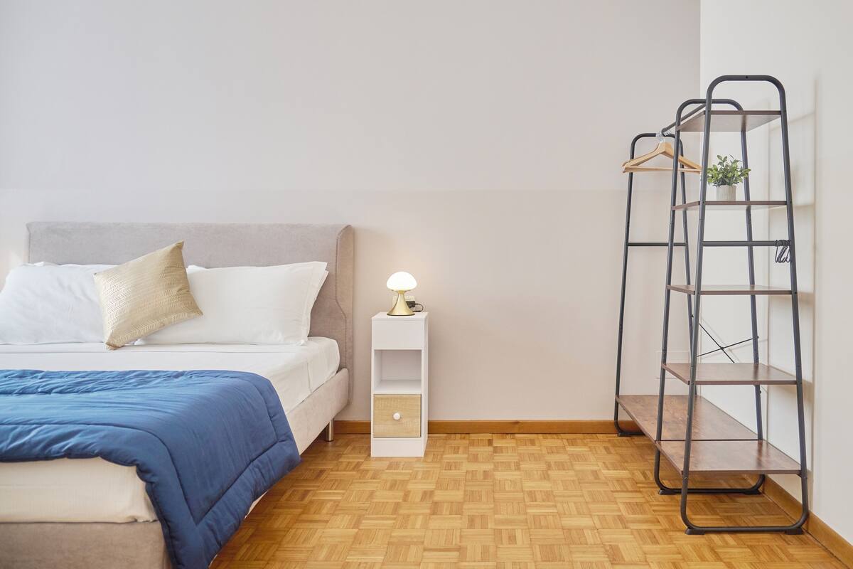 A neatly arranged bedroom with a bed, bedside table, and a metal shelf unit against the wall.