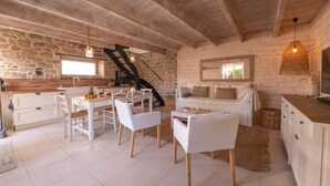 Un intérieur rustique avec un mur en pierre, des poutres en bois au plafond, un coin cuisine, des meubles blancs et un escalier.