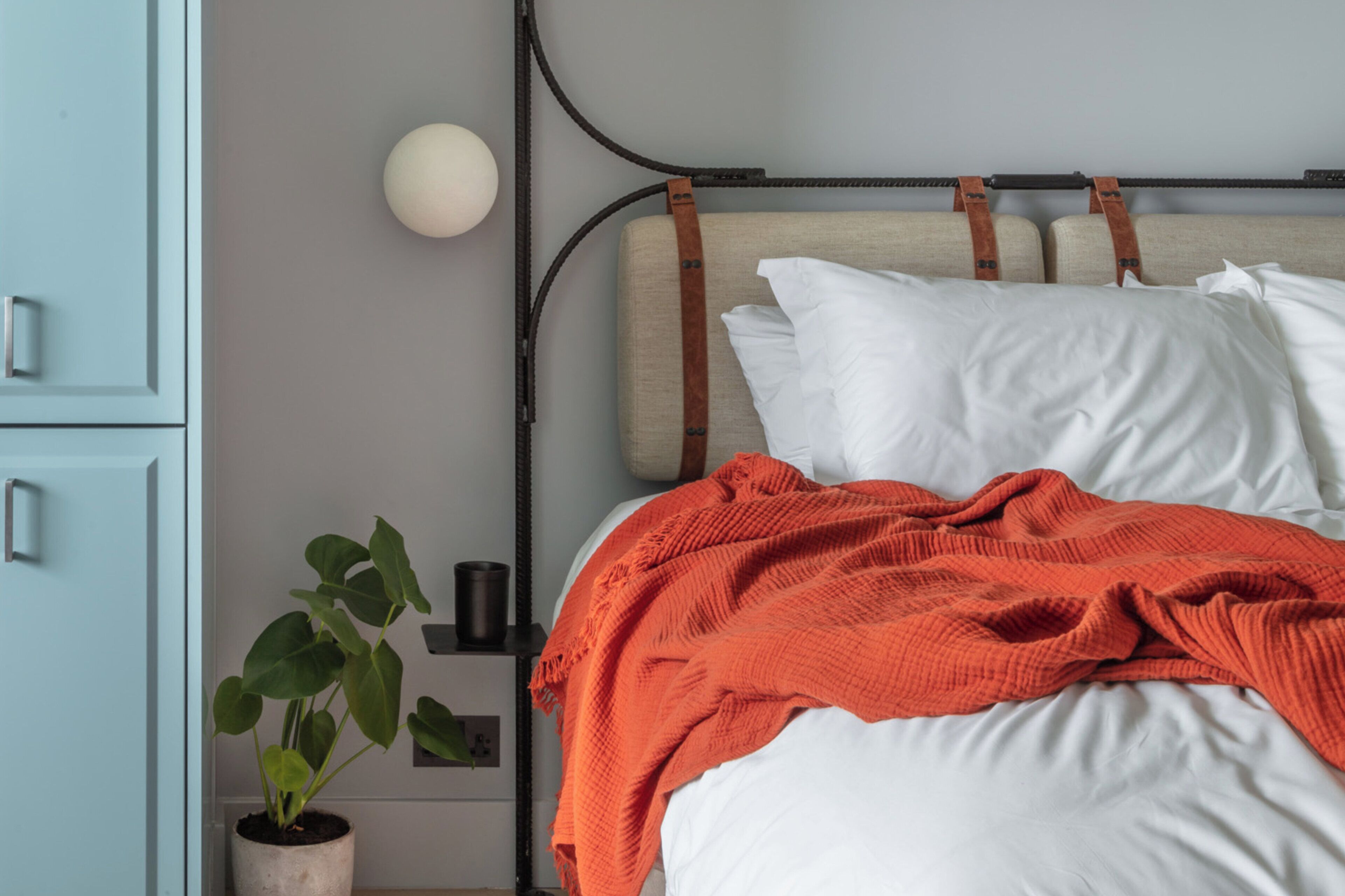 Una cama con ropa de cama blanca y una manta naranja, una planta en la mesita de noche, una lámpara y un aparador azul.