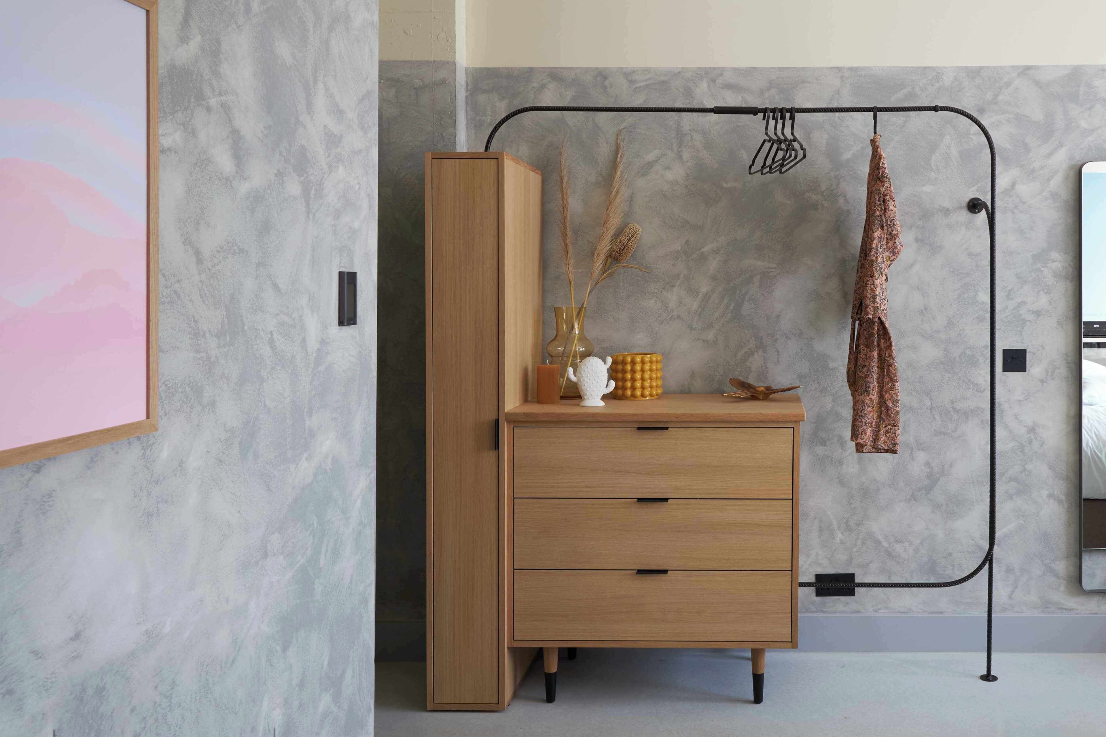 A room with a wooden dresser, a metal rack with hanging items, and a framed picture on the wall.