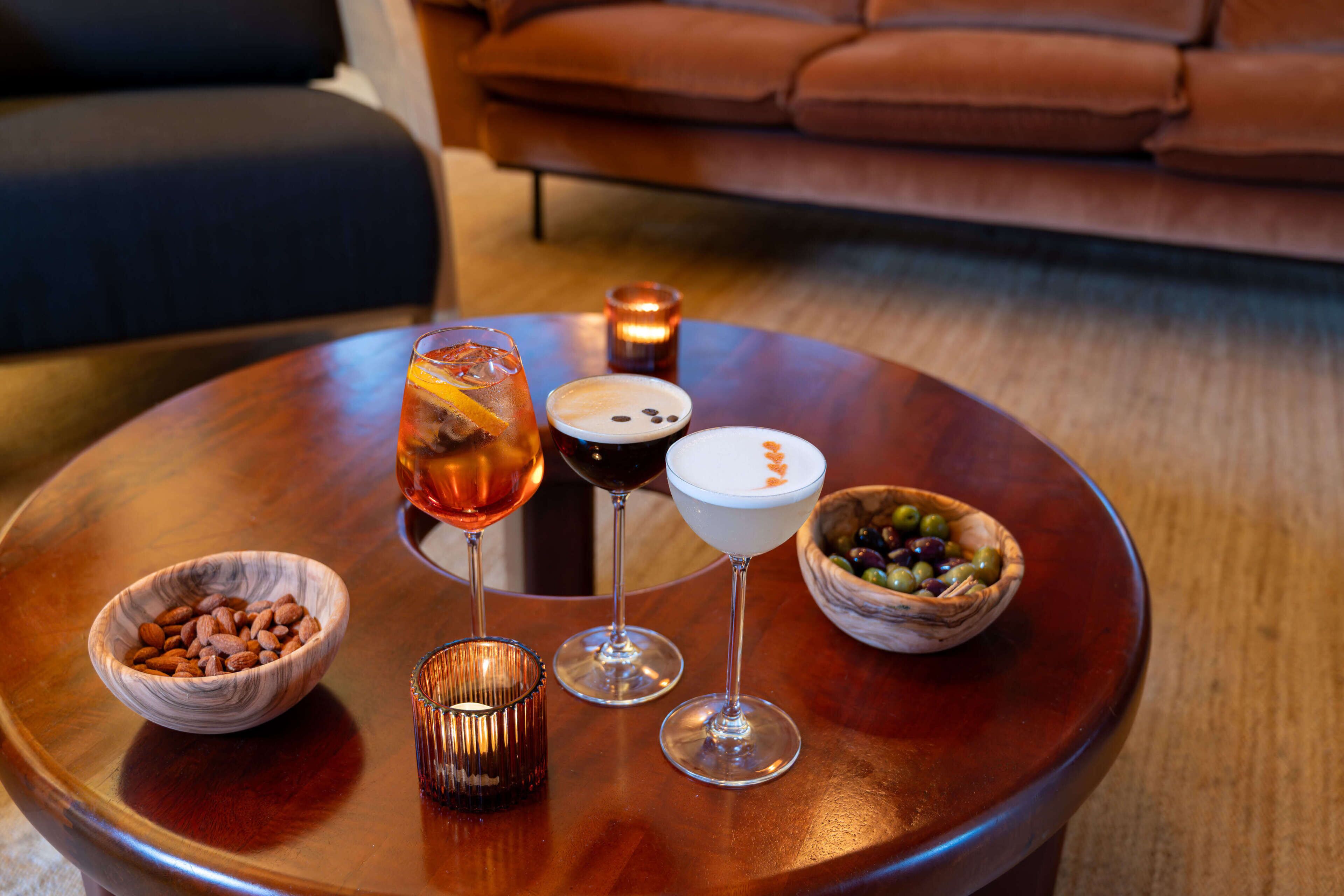 A round wooden table with three glasses of beverages, two bowls of nuts, and a candle.