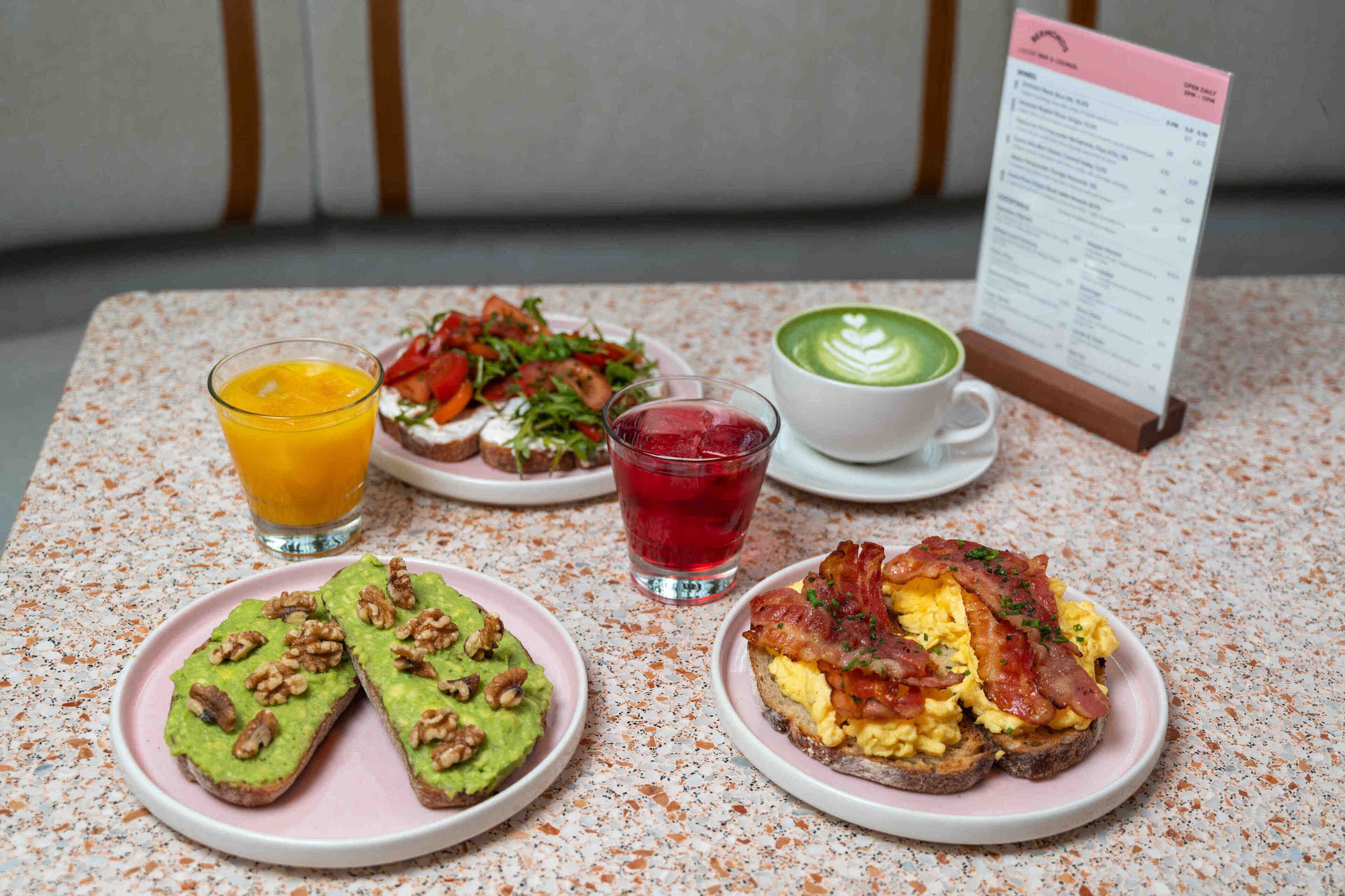 A breakfast setting with avocado toast, scrambled eggs with bacon, a green smoothie, and a red drink on a table.