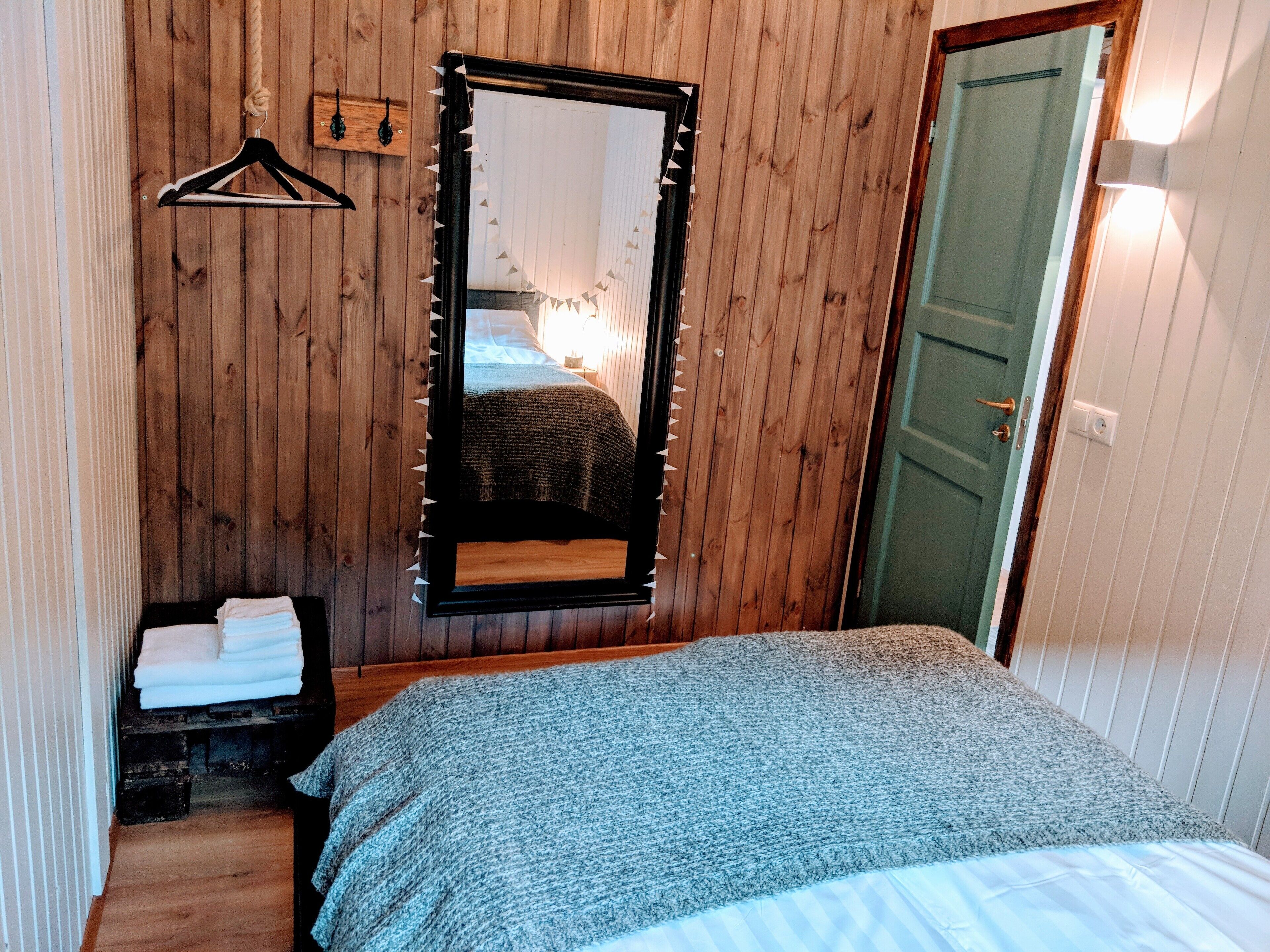 A bedroom with a wooden wall, a bed with a grey blanket, a hanging mirror, and a door.