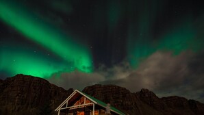A wooden cabin with a sloped roof, surrounded by mountains, under a night sky with green aurora borealis. - Múlakot Cosy Cabins (Borgarbyggd)
