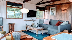 A cozy living room with a wooden ceiling, a grey sofa, a wooden coffee table, and a large window with a view of greenery. - Múlakot Cosy Cabins (Borgarbyggd)