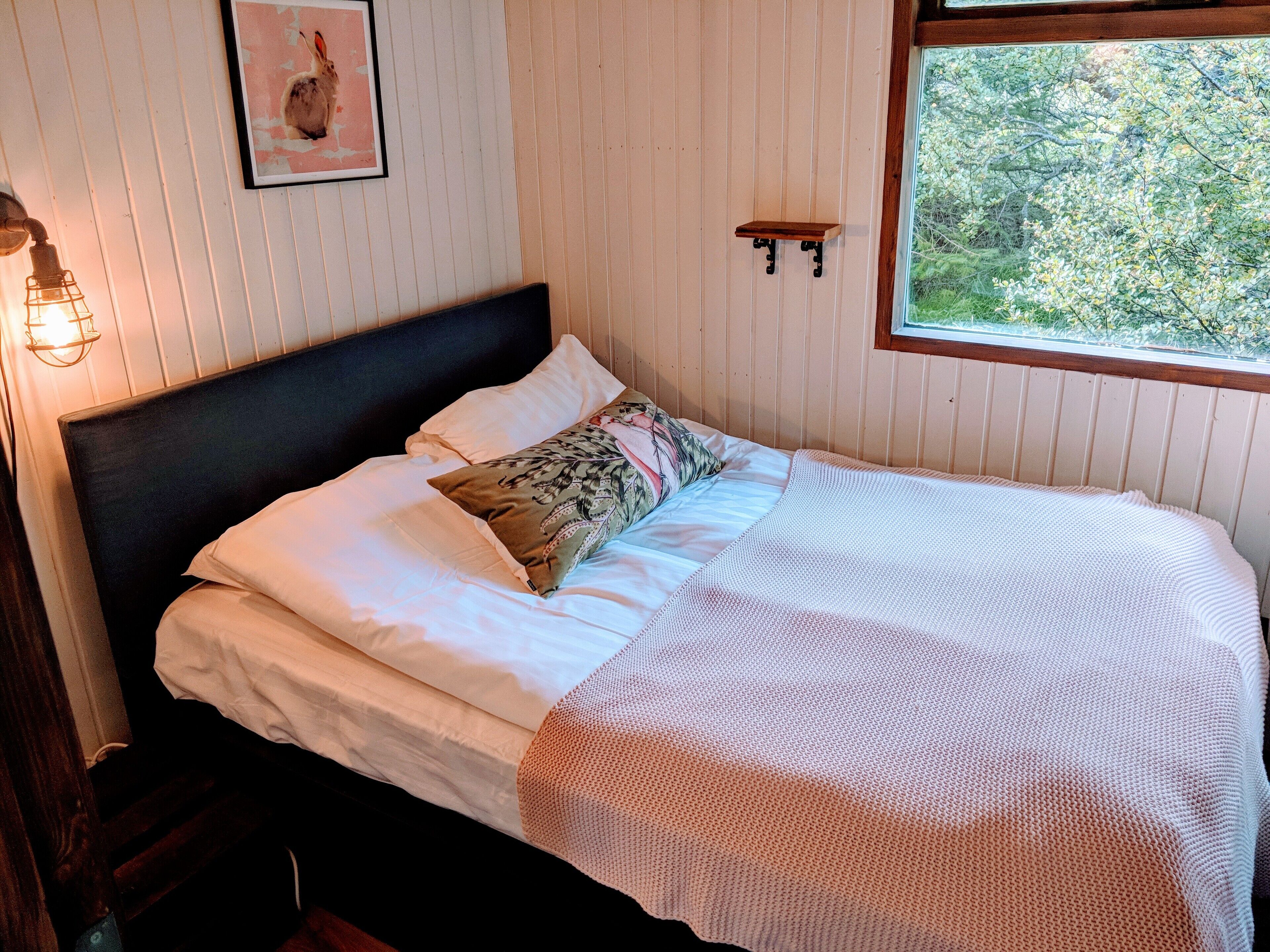 Een net opgemaakt bed met een witte dekbedovertrek en een kussen met dessin, een ingelijste foto aan de muur en een klein plankje met een plantje.