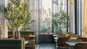 A modern interior with a long corridor lined with tables and chairs, large windows offering a view of the city, and hanging plants adding greenery. - The Glenmark, Glendale, a Tribute Portfolio Hotel (Glendale)