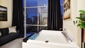 Een moderne badkamer met een badkuip, een raam met glaswand, een leren bank in zwart leer en een stadszicht in de schemering.