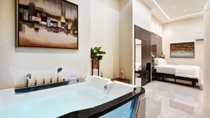 Een moderne badkamer met een groot bad, een douche en een toilet. Er hangt een schilderij aan de muur en in de aangrenzende kamer staat een bed.