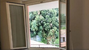 Ein Zimmer mit großem Fenster und Blick auf dichtes Grün sowie eine Steinmauer.