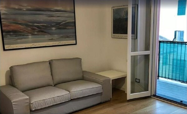 A living room with a grey sofa, a small table, and a sliding glass door leading to a balcony.