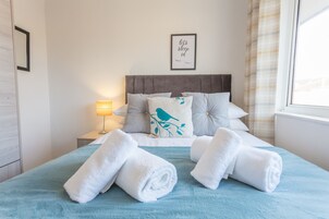 A neatly made bed with a blue blanket, white pillows, and a bird-themed pillow. There are rolled white towels on the bed, a bedside lamp, and a framed quote on the wall.