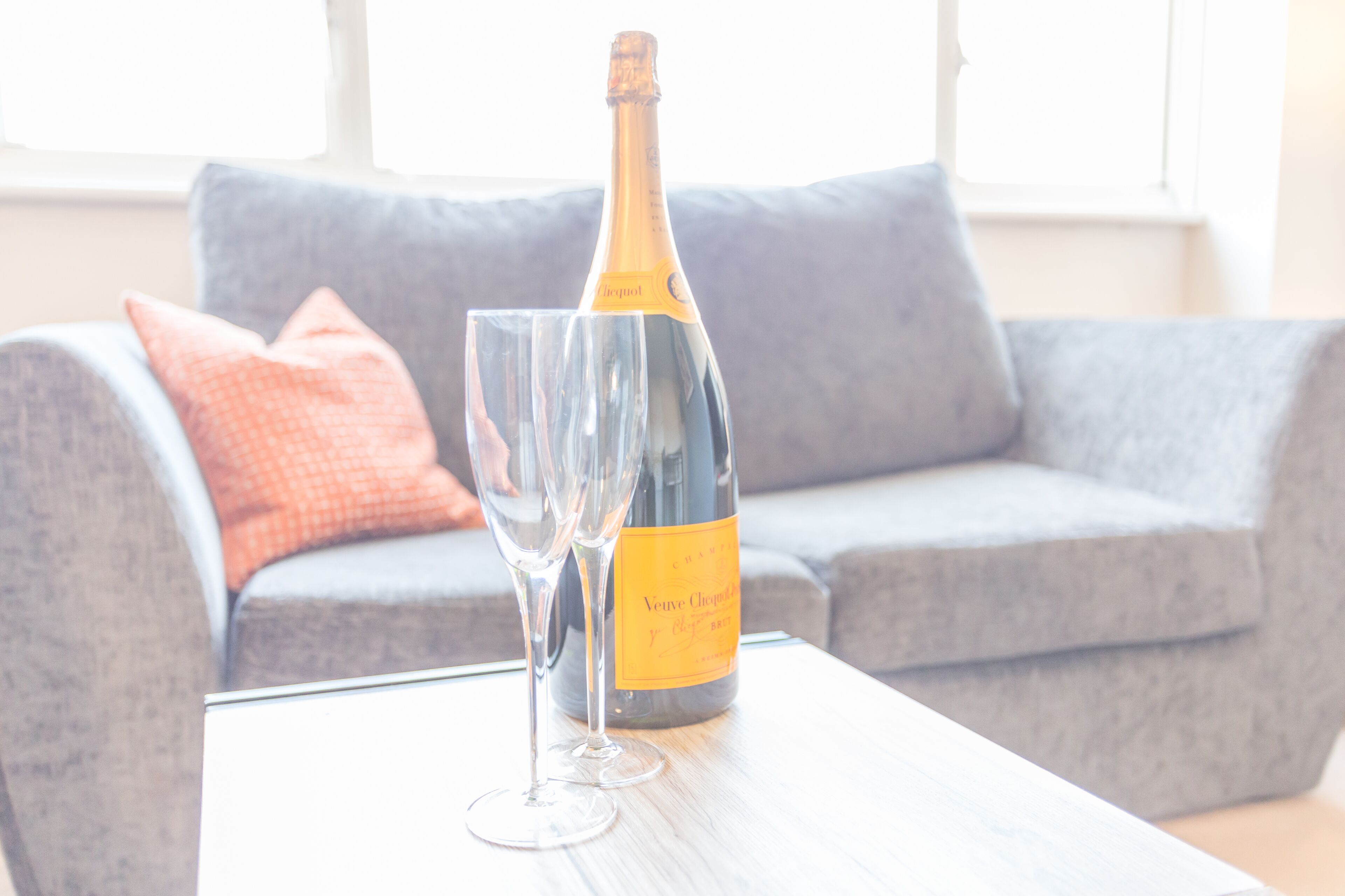 A bottle of Veuve Clicquot champagne and two glasses on a table in a living room with a grey sofa and a window in the background.