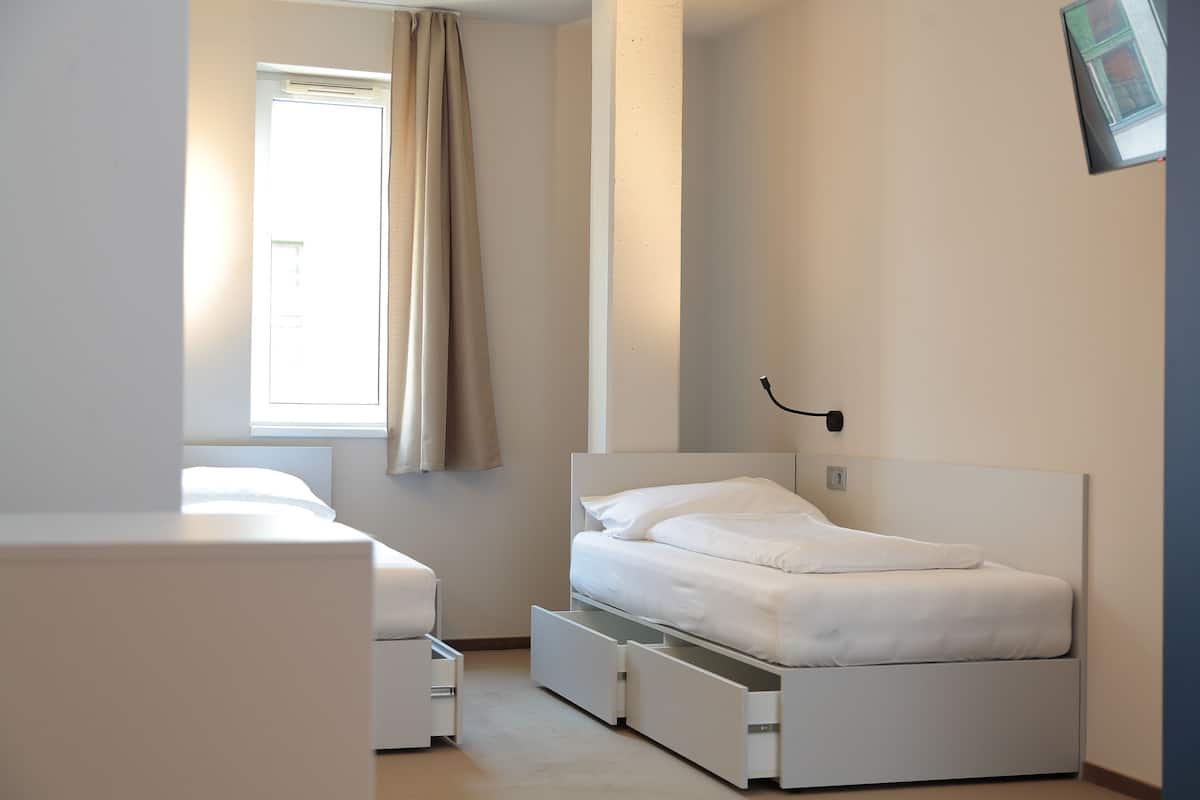 Une chambre à coucher comprenant un lit, une table de chevet, une fenêtre avec des rideaux et une lampe fixée au mur.
