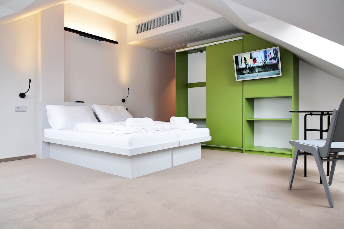 Une chambre moderne avec un lit blanc, des murs d’un vert vif, un téléviseur fixé au mur et un fauteuil.