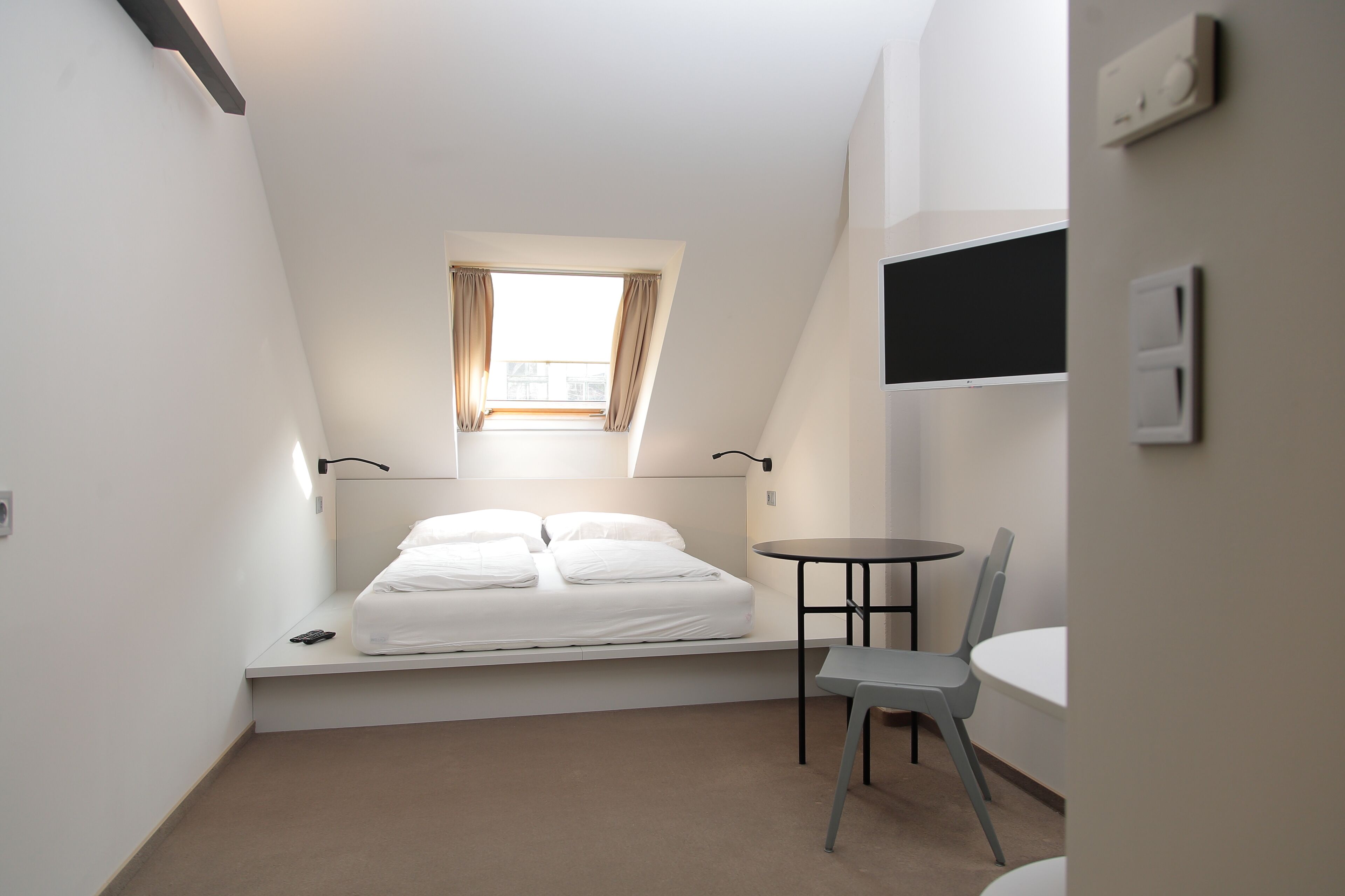 Une petite pièce bien éclairée, avec un lit, une chaise, une petite table et une fenêtre avec des rideaux.