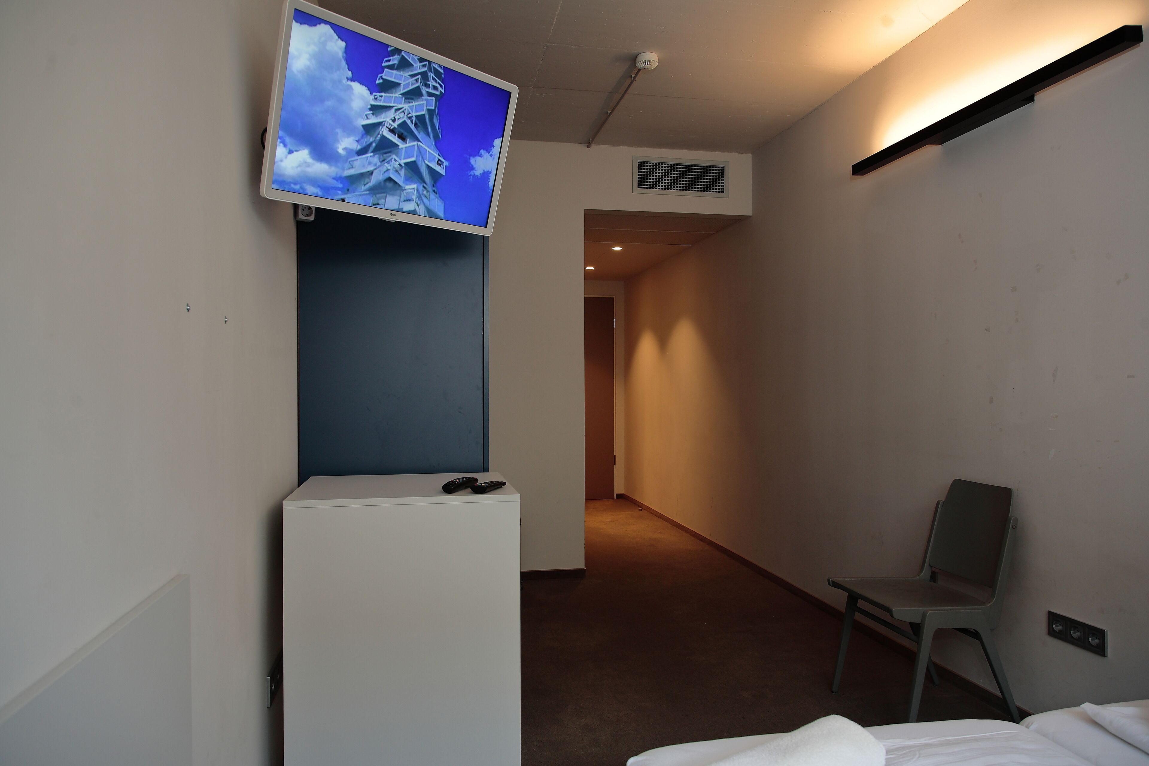 A hotel room with a flat-screen TV mounted on the wall, a small white cabinet, a grey chair, and a bed with white bedding.