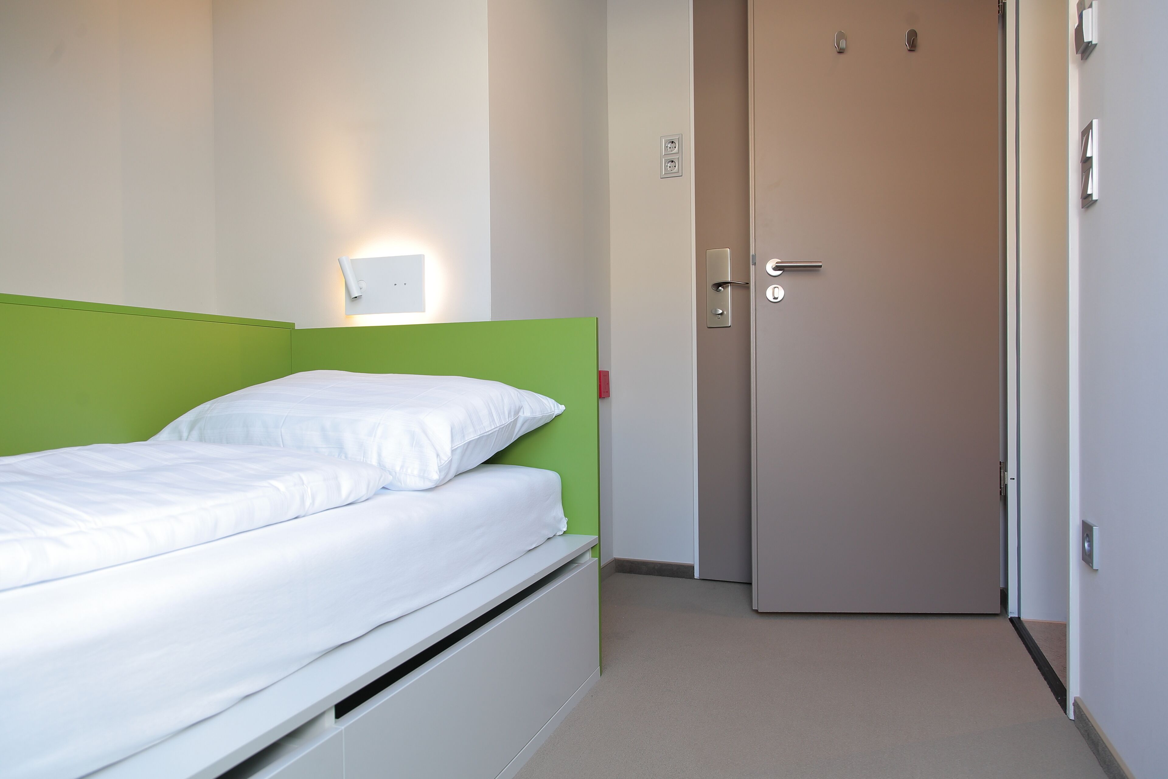 Habitación individual con una cama y cabecera verde, una mesita de noche y un interruptor de luz en la pared.