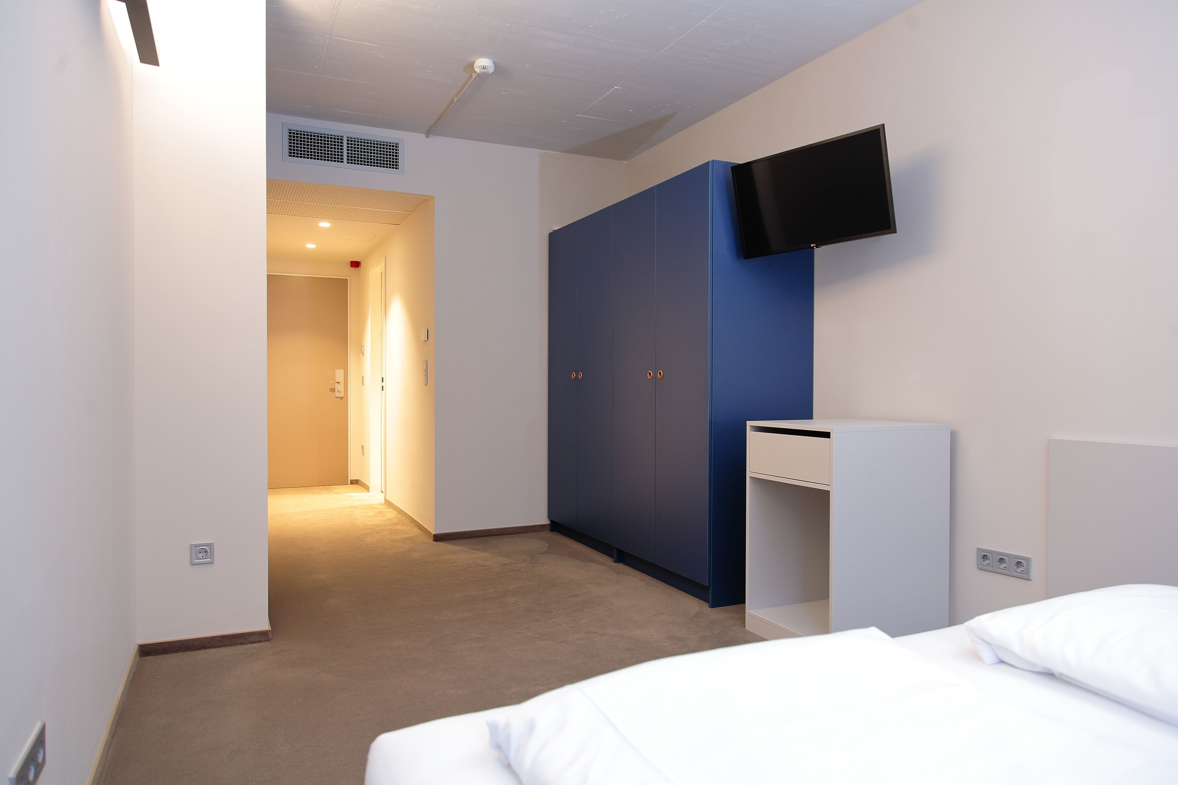 Habitación de hotel con cama, armario, televisor montado en la pared y un pasillo que conduce a otra habitación.