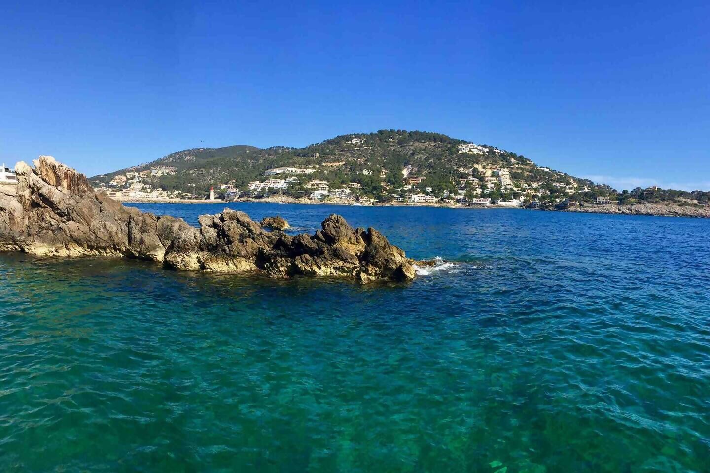 A coastal landscape with clear blue waters, rocky formations, and a hill with buildings in the background.