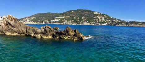 A coastal landscape with clear blue waters, rocky formations, and a hill with buildings in the background.