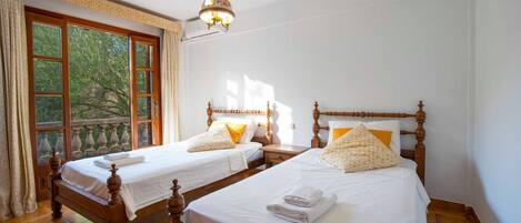 A room with two wooden beds, a balcony with a view of trees, and a hanging light fixture.