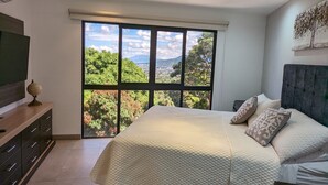 Ein Schlafzimmer mit großem Fenster, einem Bett mit gesteppter Bettdecke, einem Nachttisch und einem Fernseher auf einem Schrank.