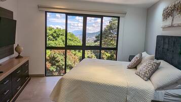 Un dormitorio con un ventanal grande, una cama con cubrecama acolchado, una mesita de noche y un televisor sobre un mueble.