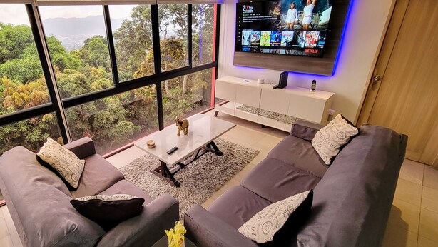 Ein modernes Wohnzimmer mit einem großen Fenster, das Blick auf üppiges Grün und Berge bietet. Es gibt ein graues Sofas mit gemusterten Kissen, einen kleinen Couchtisch und einen flachbildfernseher an der Wand, der einen Film zeigt.