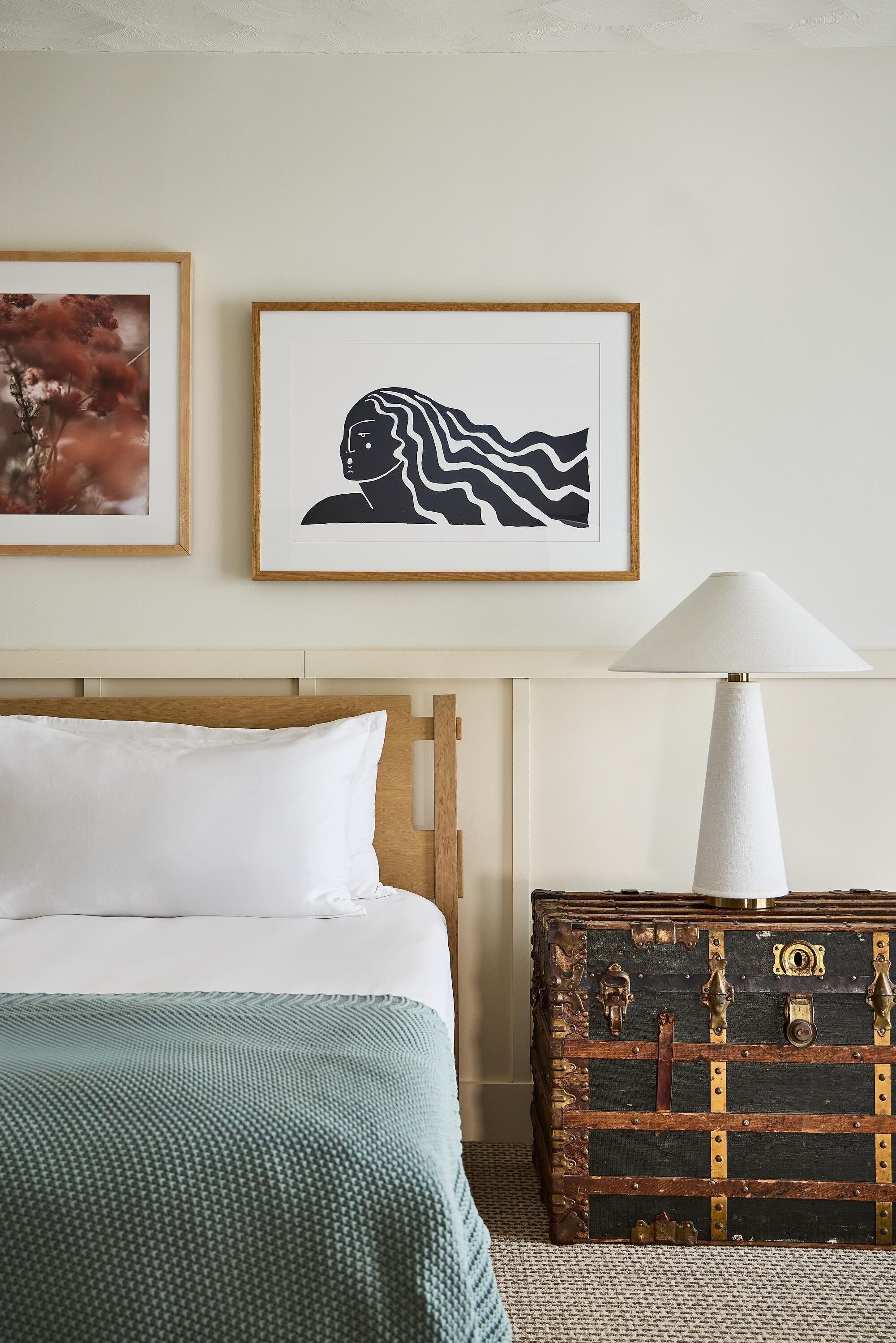 Una cama bien tendida con ropa blanca y una manta verde, una obra de arte enmarcada encima, una lámpara de noche y un baúl antiguo con herrajes de latón.