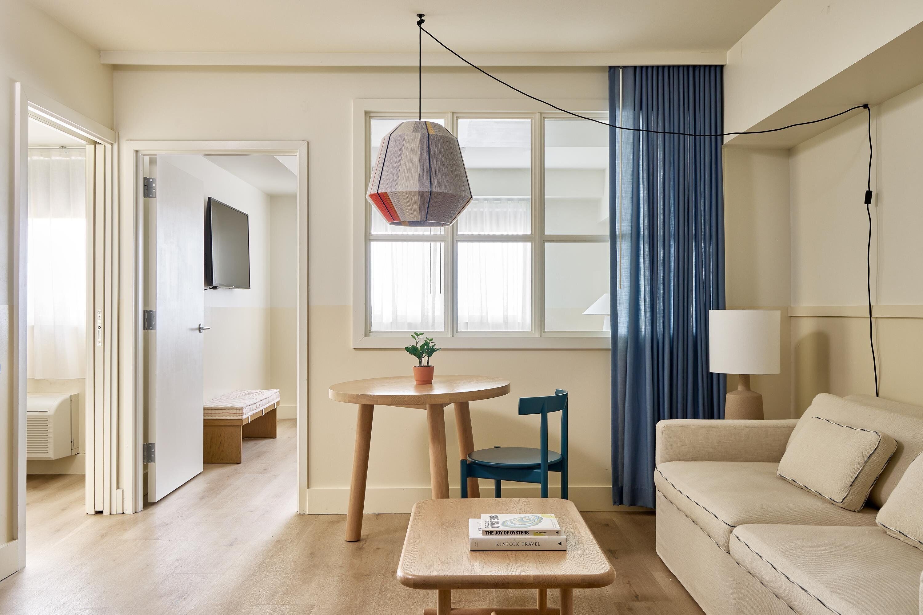 A modern living room with a sofa, a coffee table with books, a small table with a plant, a blue chair, and a large window with curtains.