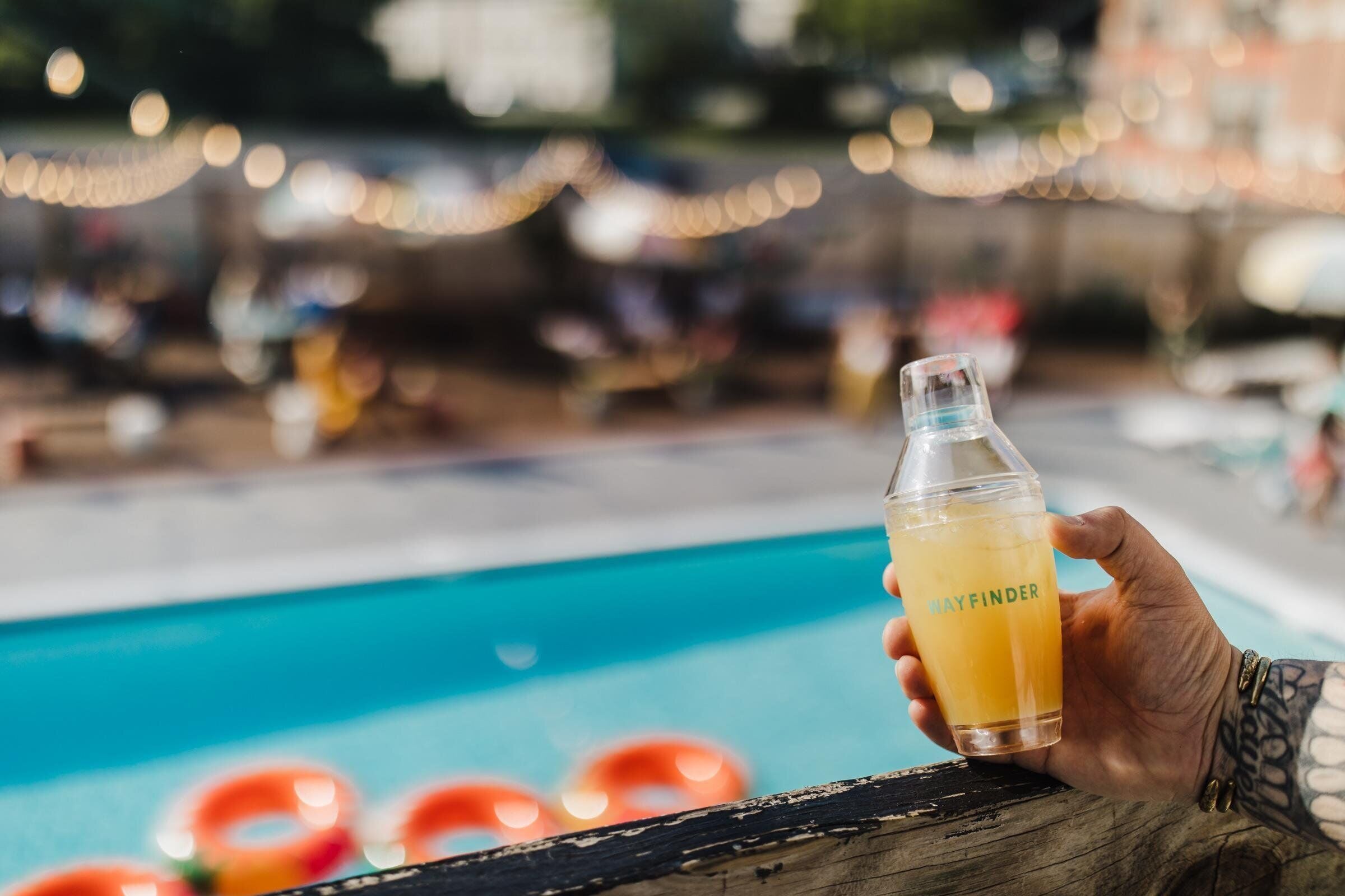 A hand holding a bottle with a label, overlooking a pool with a blurred background.