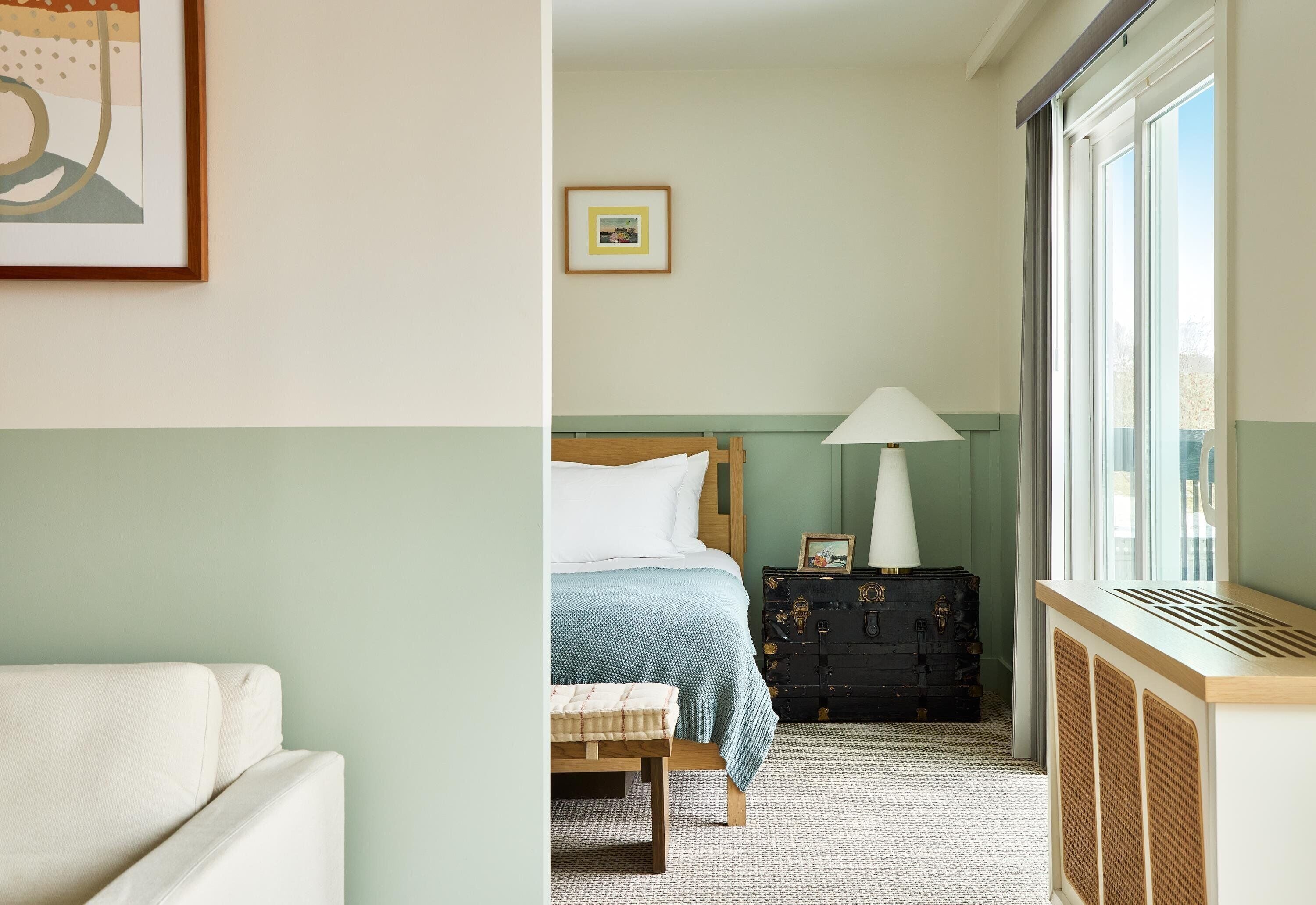 A hotel room with a bed, a bedside table with a lamp, a suitcase, and a window with a view.