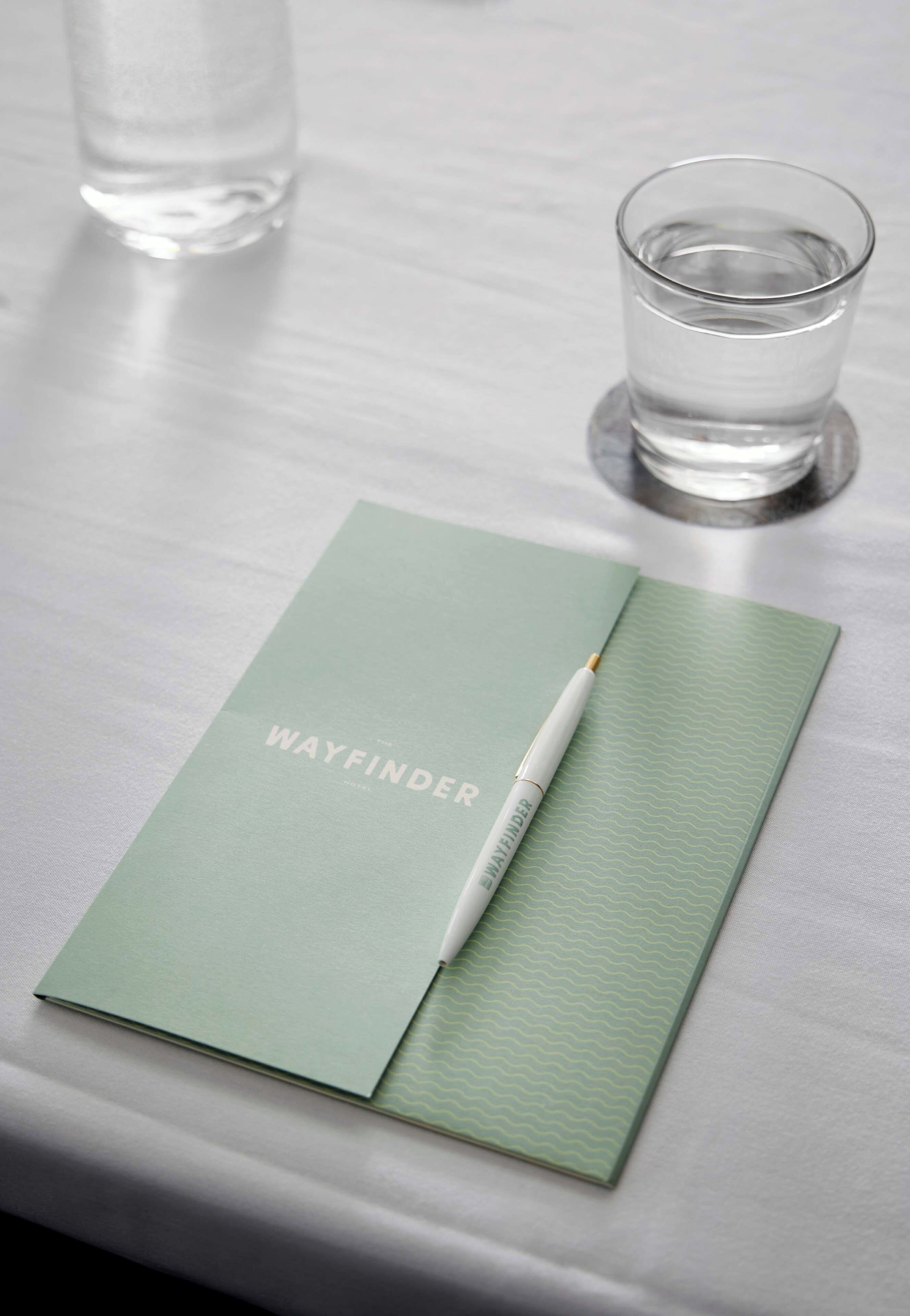 En notesbog med teksten 'WAYFINDER' ligger på et bord sammen med en pen og et glas vand.