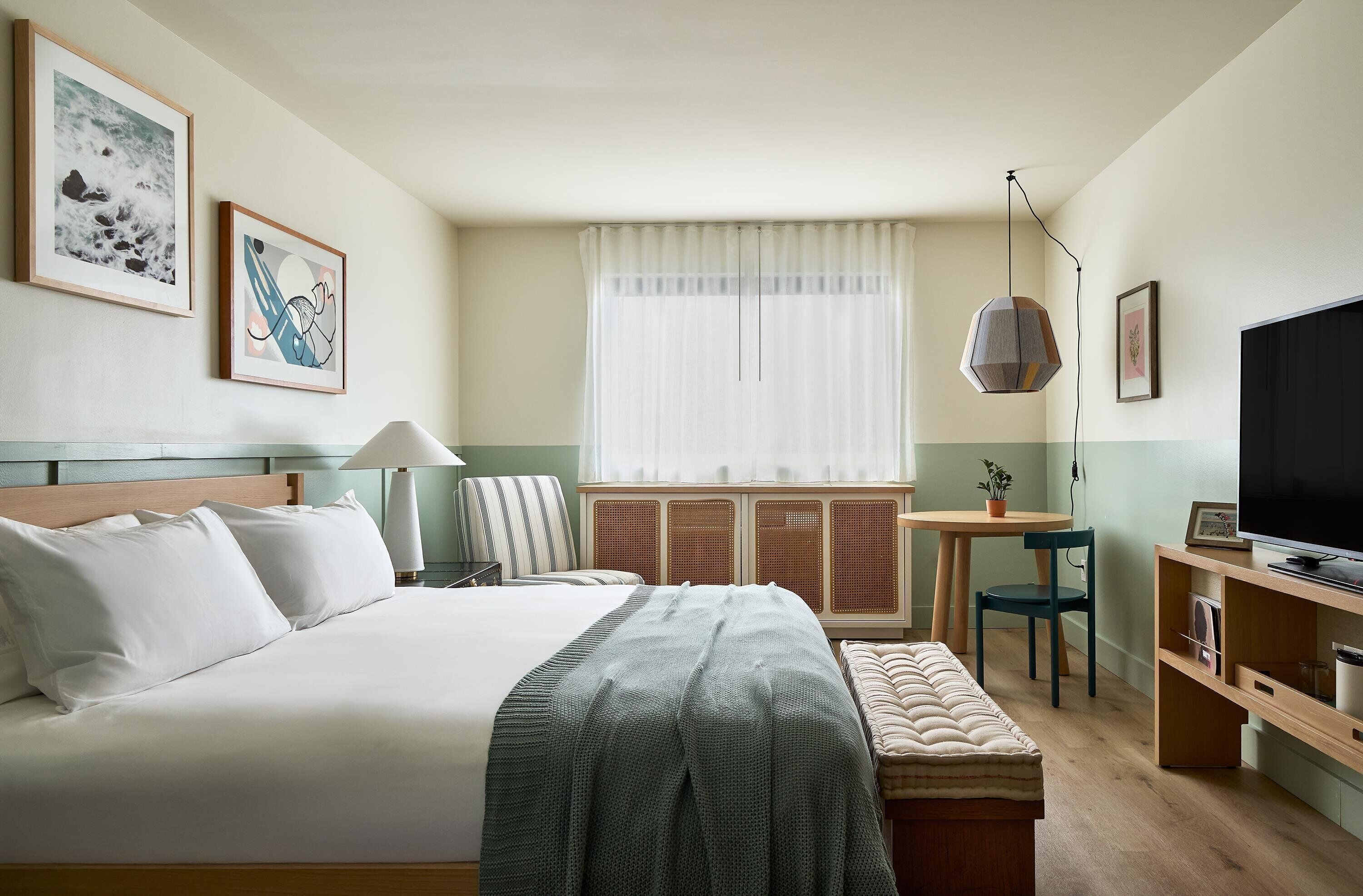 Quarto de hotel com cama, mesa de cabeceira, luminária, poltrona, televisão e uma janela com cortinas.
