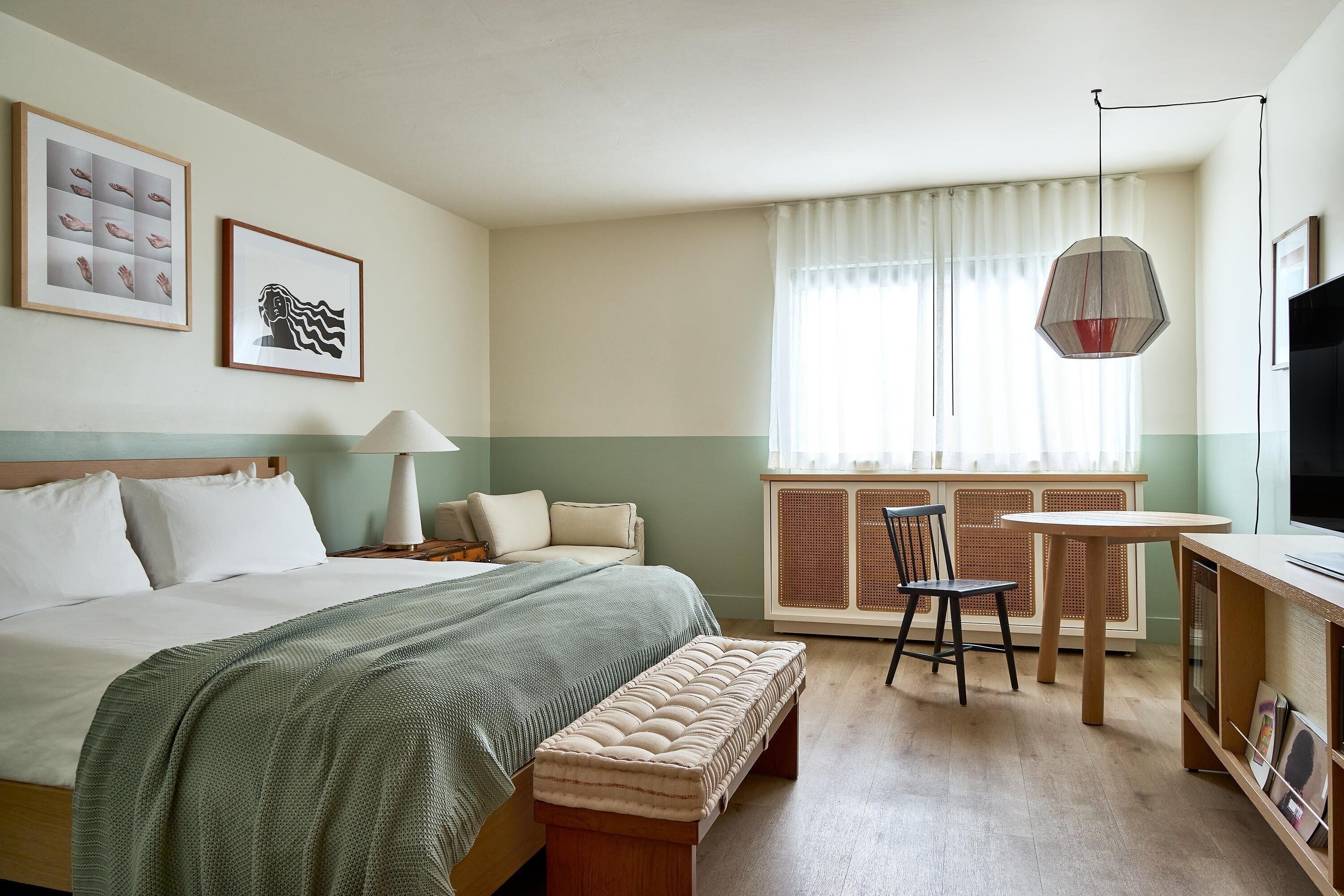 Habitación de hotel con cama, escritorio, silla, lámpara, televisor y obras de arte en la pared.