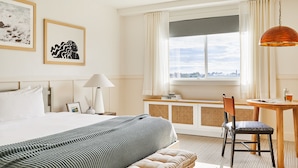 A hotel room with a large bed, a desk with a chair, a window with curtains, and artwork on the wall.