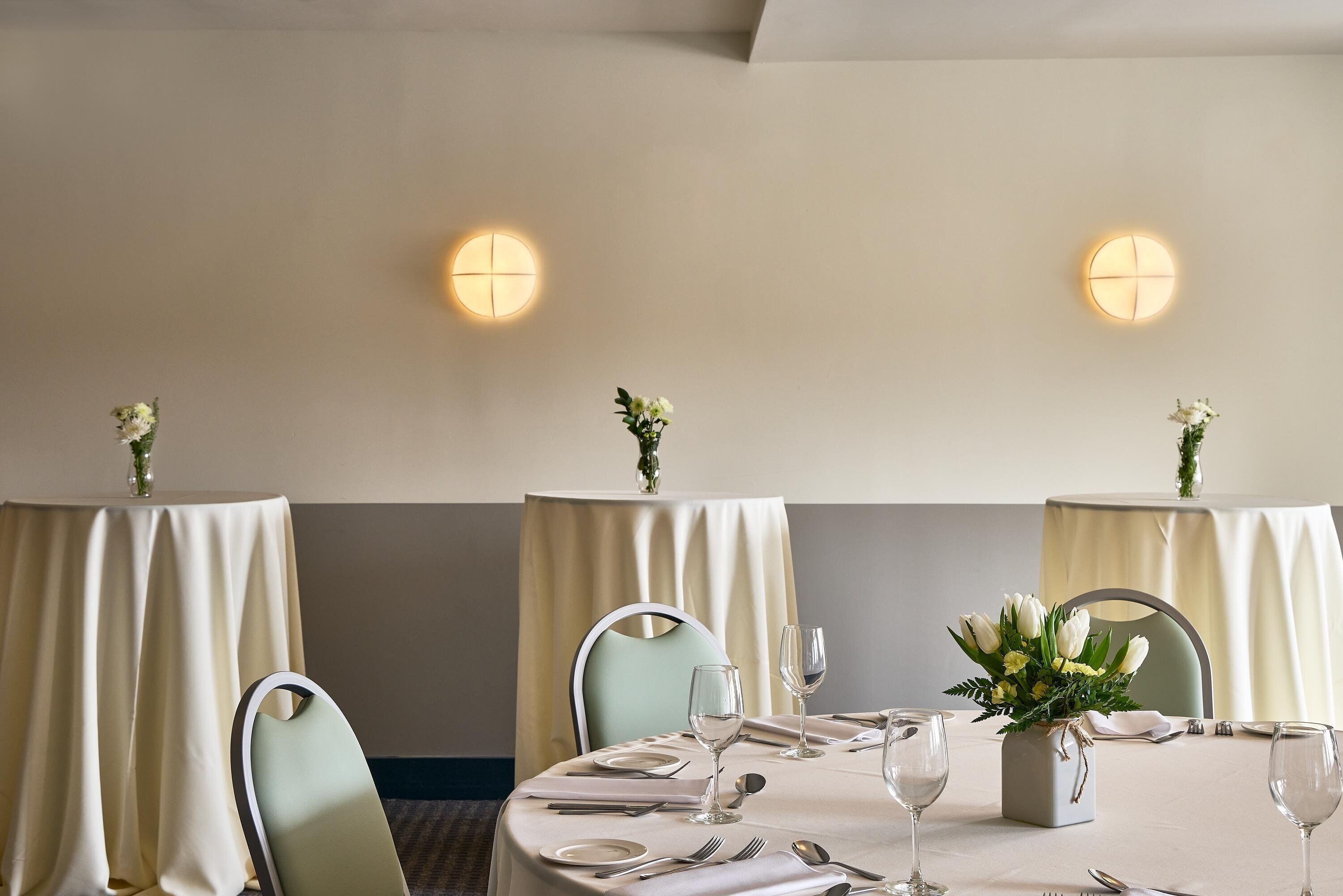Un comedor con mesas redondas, sillas y lámparas fijadas a la pared.