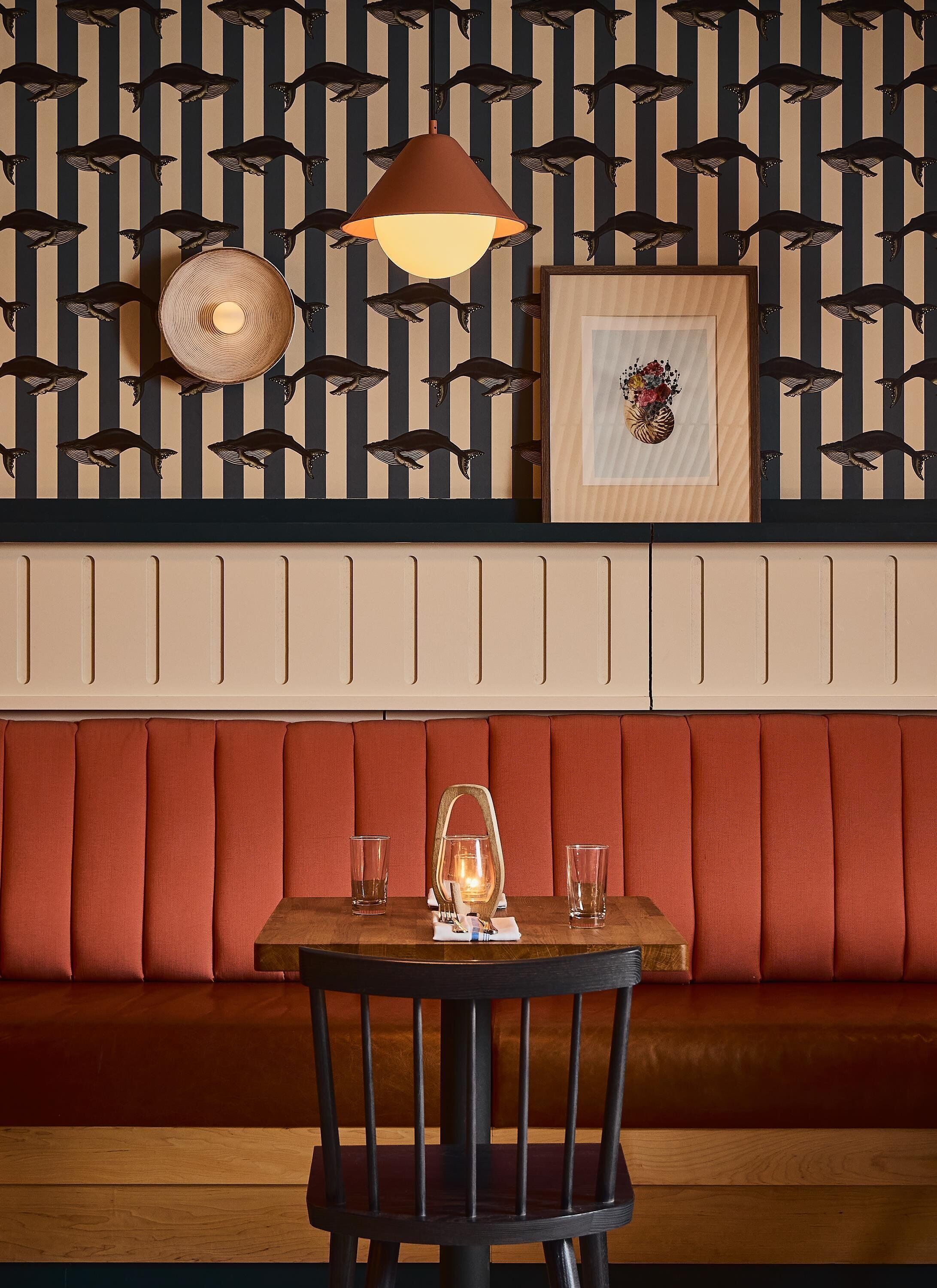 A dining area with a wooden table, a black chair, a red cushioned bench, a hanging pendant light, and a wall with a striped pattern.
