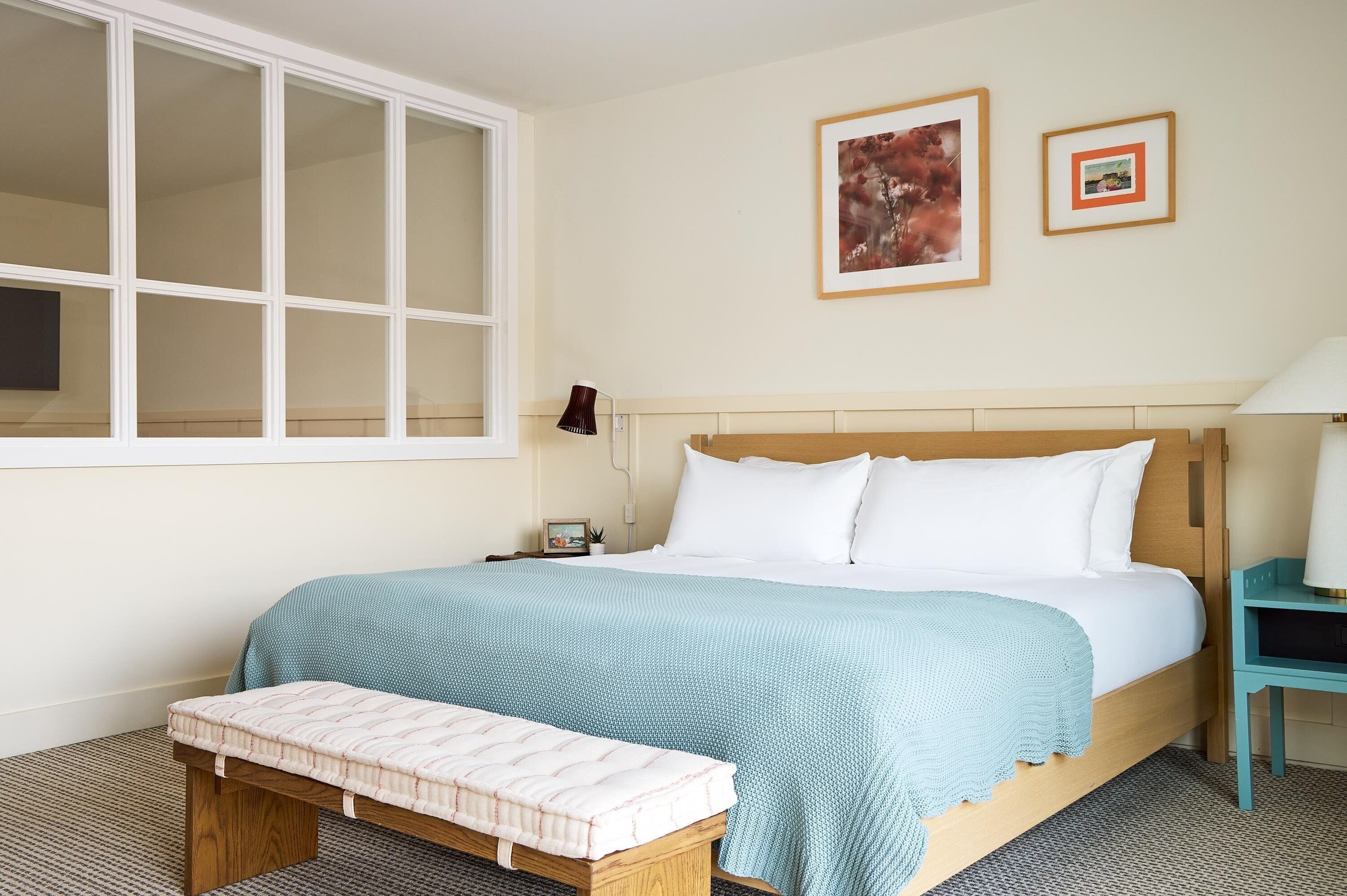 A bedroom with a large window, a bed with white bedding, a bedside table with a lamp, and two framed pictures on the wall.
