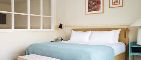 A bedroom with a large window, a bed with white bedding, a bedside table with a lamp, and two framed pictures on the wall.