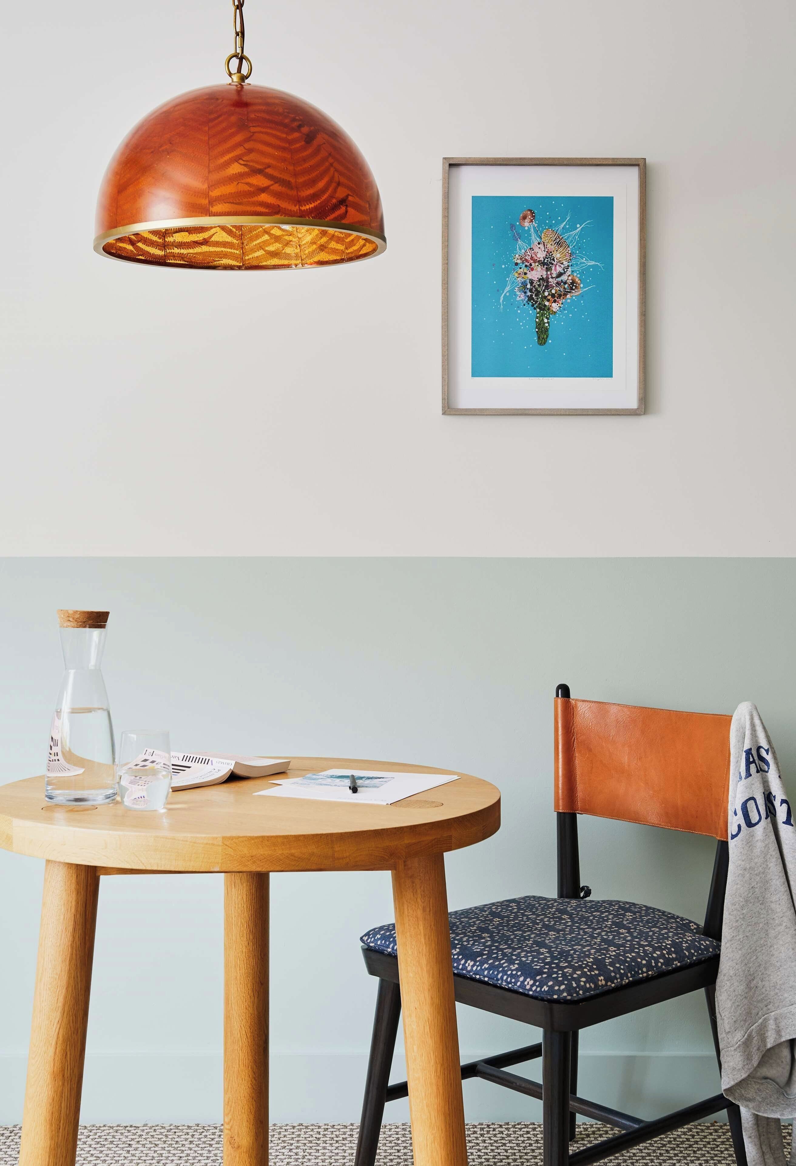 Une table ronde en bois, sur laquelle se trouvent une bouteille en verre transparent, un verre et des papiers. Une chaise avec un coussin à motifs et une serviette drapée sur le dossier est placée à côté de la table. Au-dessus, une suspension aux finitions en bois est suspendue au plafond, et un tableau abstrait encadré est accroché au mur.