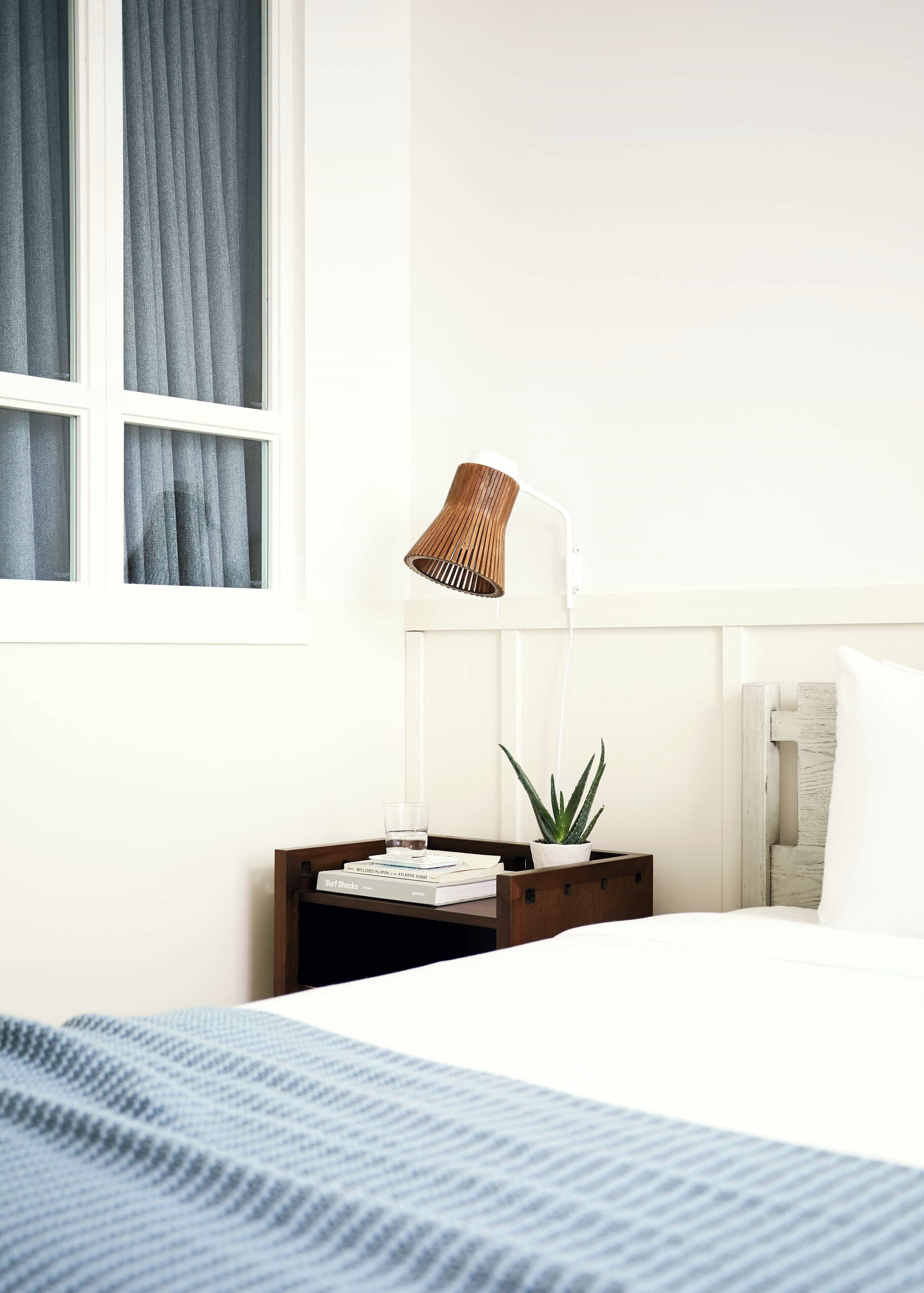 Un lit bien fait, recouvert d’une couverture bleue, une table de chevet avec une lampe, une plante en pot et une pile de livres.
