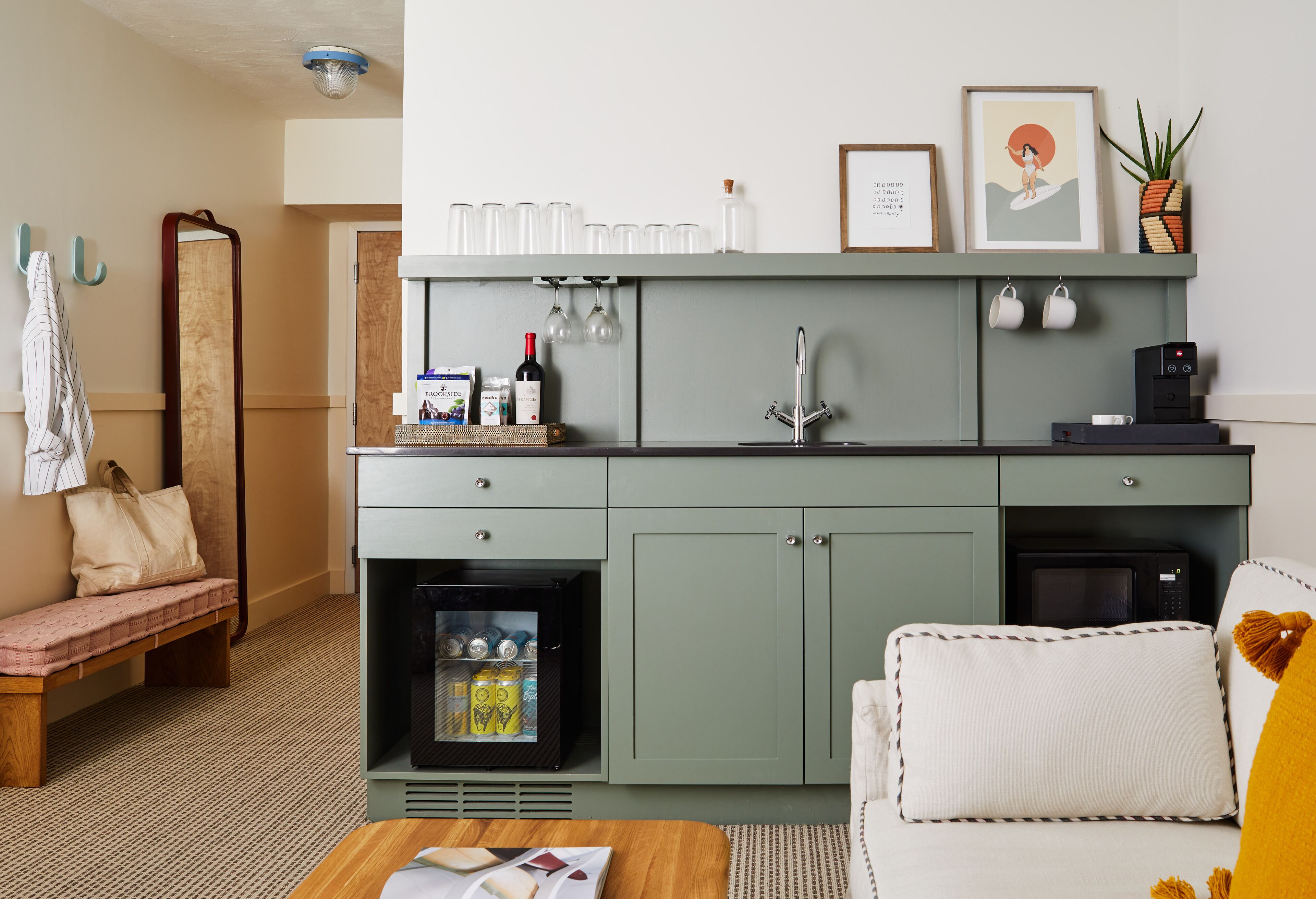 A compact kitchenette with a fridge, microwave, and a small coffee machine. There is a bench and a sofa in the room.