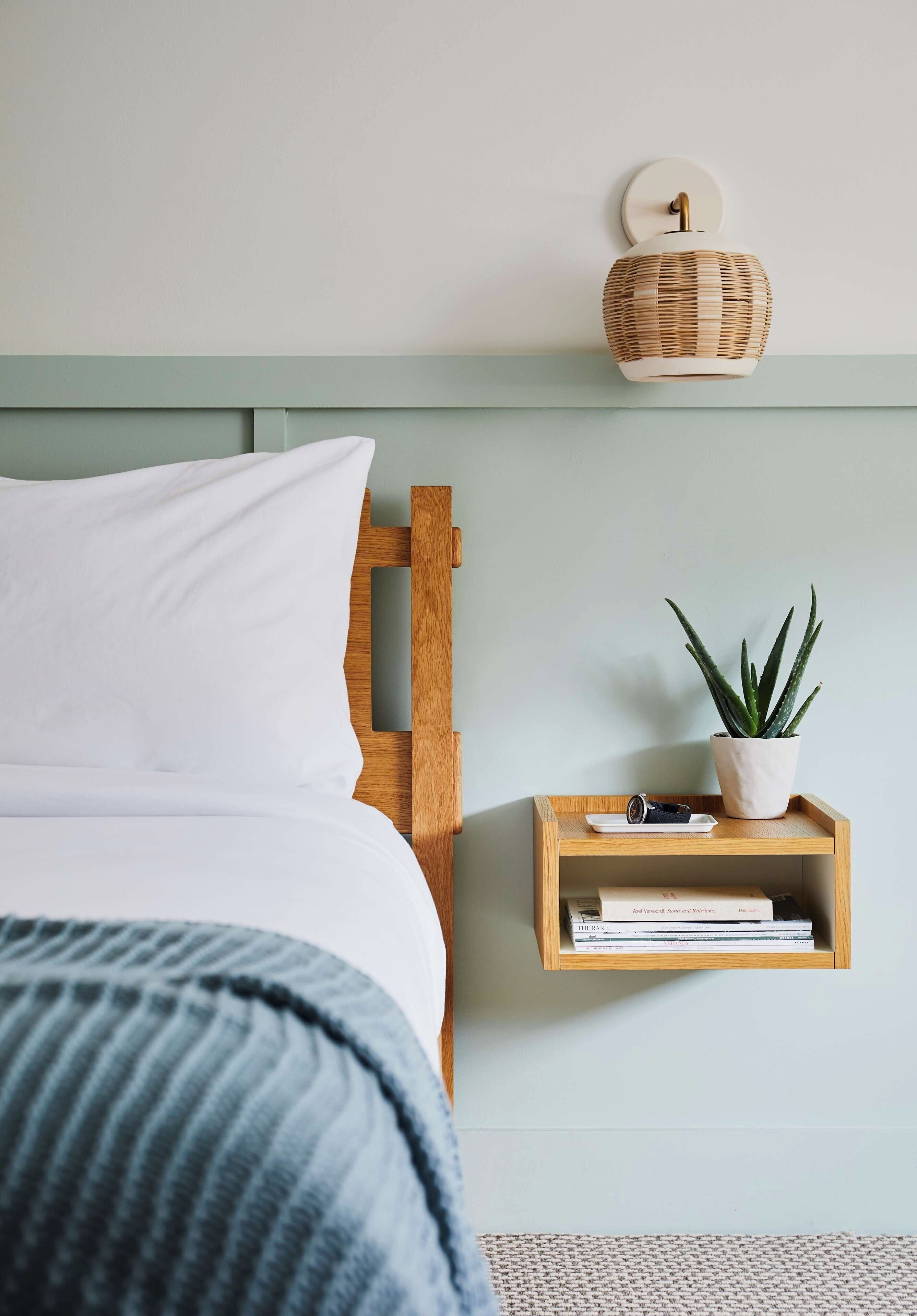 Une chambre à coucher avec un lit, une table de chevet ornée d’une plante et de livres, et une lampe fixée au mur.