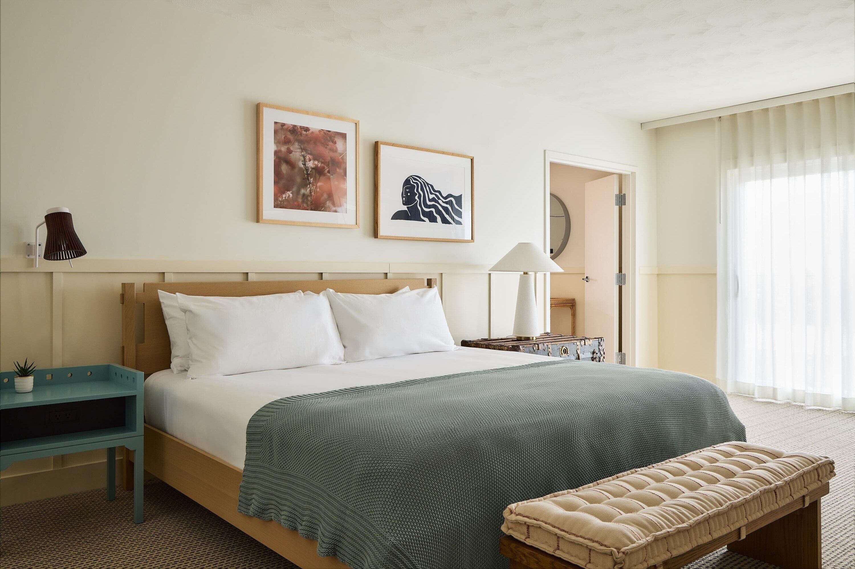 Een hotelkamer met een bed, een nachtkastje, een lamp, een bankje en twee ingelijste kunstwerken aan de muur.
